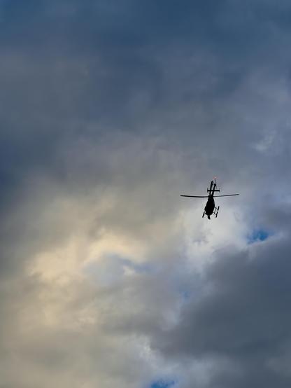 A color, wide-angle, low-angle photograph in portrait orientation that captures the dark silhouette of a helicopter flying through a dramatic, cloudy sky illuminated by the soft, golden glow of sunrise. The helicopter is positioned toward the lower right of the frame, angled slightly upward with its main rotor blades spread horizontally and a small red light blinking on its tail assembly. The vast expanse above and around the aircraft is filled with dense, billowing gray and blue clouds, while a break of warm, pale yellow light softly brightens the lower left area of the dramatic morning scene.