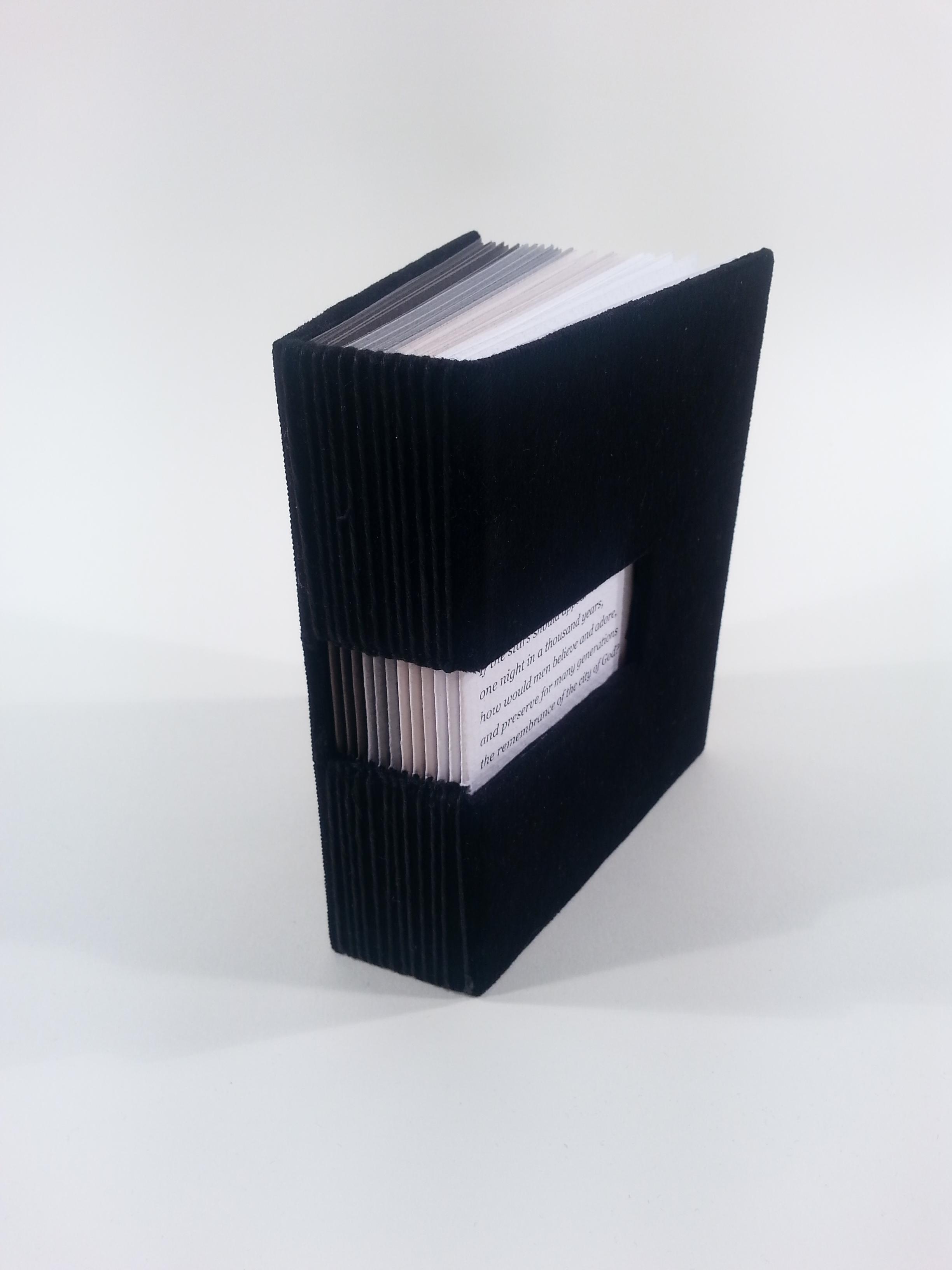 A small book with a black velvet cover and a hole in the spine that wraps around to the cover. Through the hole in the cover some text can be seen. The pages of the book progress from white to light gray to medium gray to very dark gray.