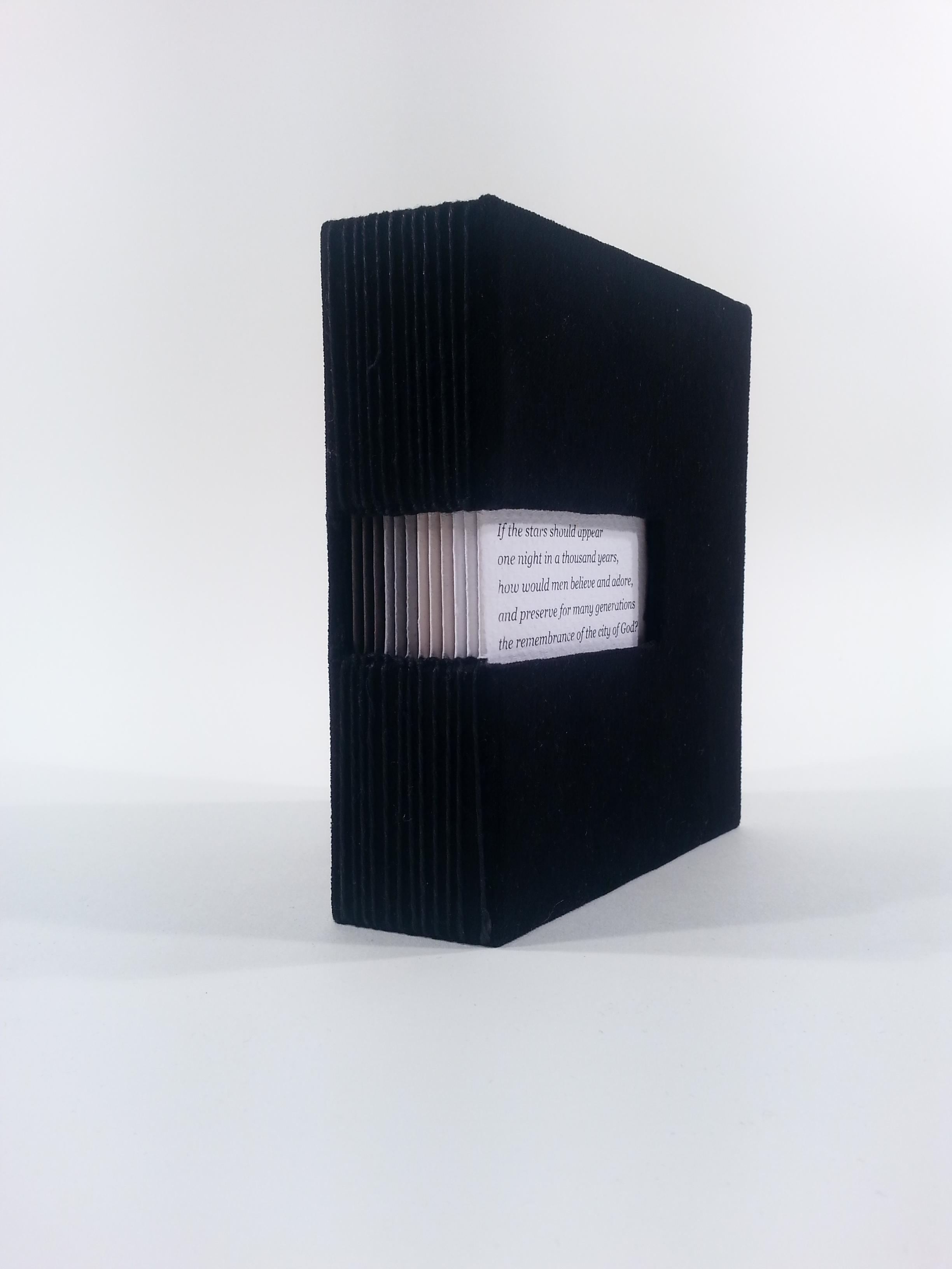 A small book with a black velvet cover and a hole in the spine that wraps around to the cover. Through the hole in the cover some text can be seen. The pages of the book progress from white to light gray to medium gray to very dark gray.