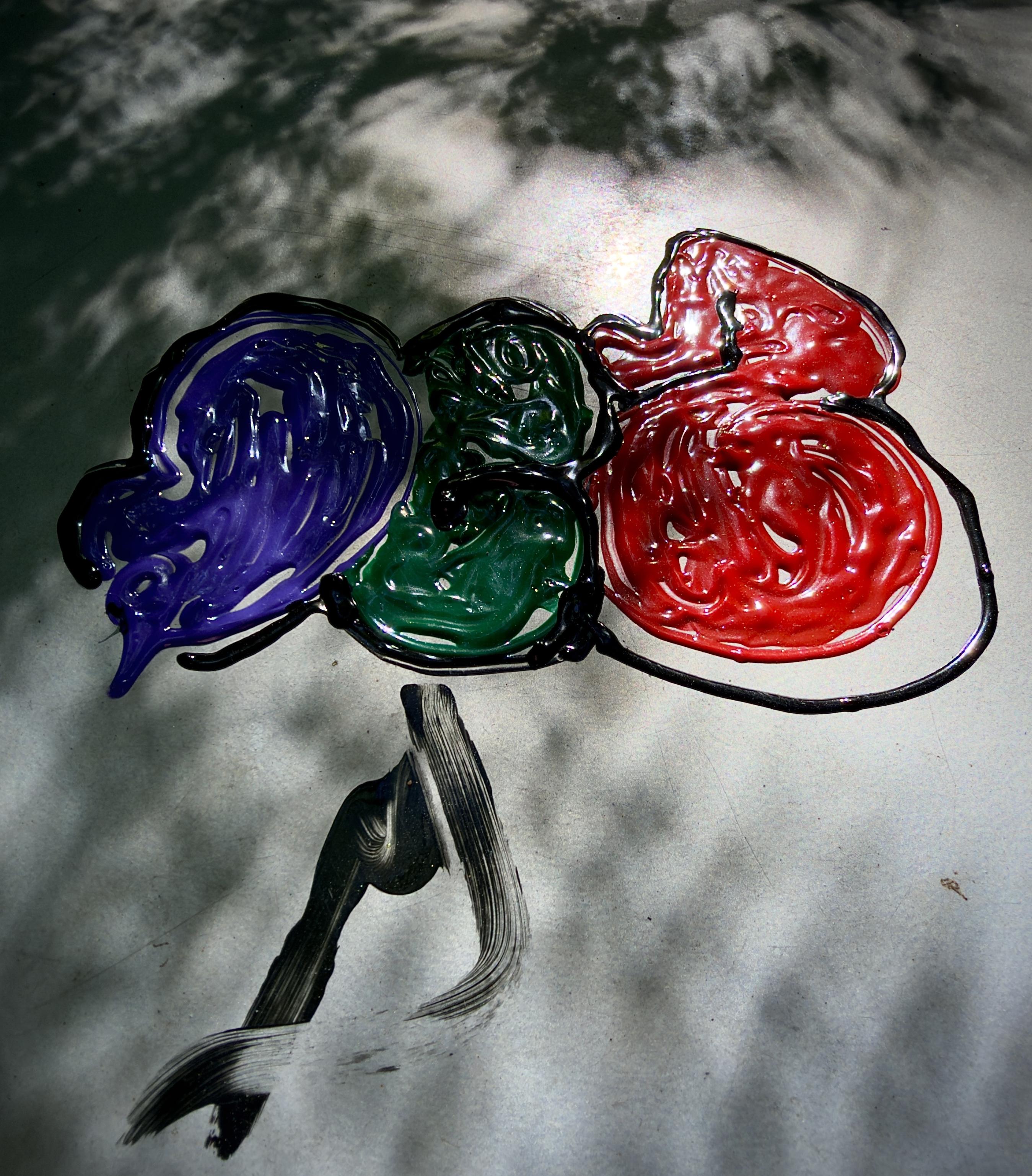 A color, medium-angle photograph in portrait orientation that captures an abstract artwork created with thick, glossy paints on a light gray, textured table along the Bowery in Manhattan. The central focus is a composition of three interconnected, bulbous shapes outlined in thick black paint, resembling fluid blobs or abstract bubbles. Each shape is filled with a distinct, vibrant color: the left section contains a deep, swirling blue, the middle section features a rich dark green, and the largest section on the right is filled with a glossy, textured red. The paint has a three-dimensional, wet appearance, catching the light with specular highlights that emphasize its viscous texture. Extending downward from the central green section is a separate, dynamic brushstroke composed of sweeping black streaks, creating a sense of movement or a dangling anchor-like element against the neutral background. Dappled shadows, cast by overhead foliage, stretch across the top and bottom edges of the surface, creating a mottled pattern of light and dark patches. The overall lighting is bright and natural, accentuating the glossy sheen of the thick pigment layers and the contrasting matte texture of the table underneath.
