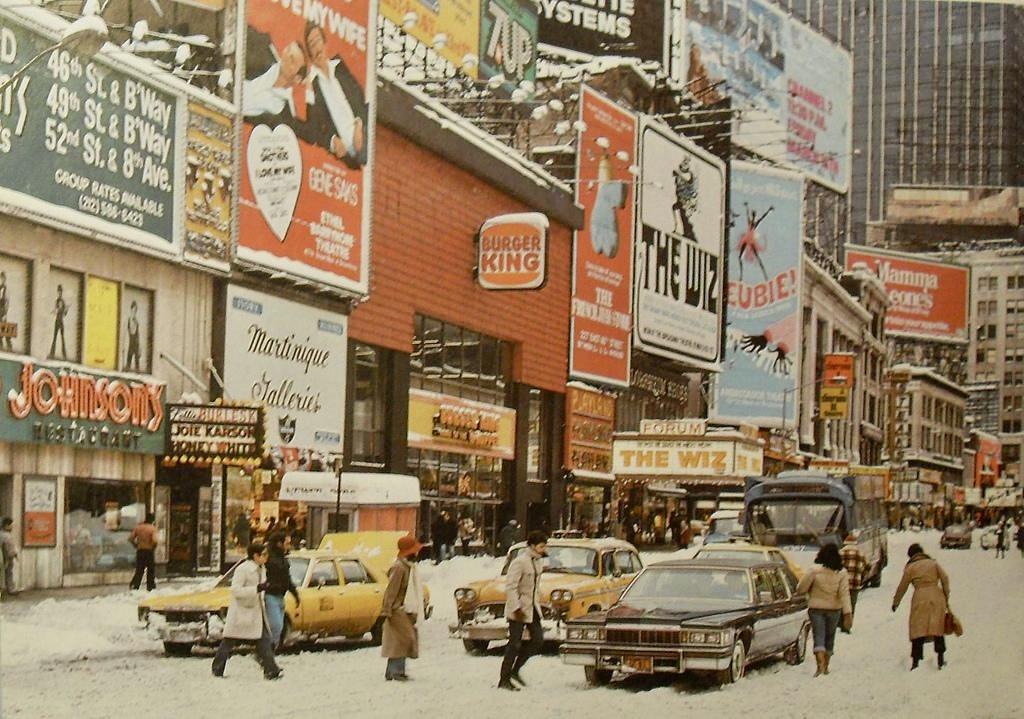A color, wide-angle photograph in landscape orientation that captures a snowy winter street scene in Times Square, Manhattan, during the blizzard of 1978. The ground and the street are completely covered in thick layers of white snow, with slushy tracks carved out where vehicles have driven. Several vintage automobiles, including a yellow taxicab on the left and a dark-colored sedan closer to the center foreground, navigate the snow-packed thoroughfare. A couple of pedestrians wearing winter coats walk along the snowy street and sidewalks, braving the cold weather while a city bus and other cars can be seen further down the avenue. The buildings lining the street are densely covered with a chaotic array of large, retro storefront signs, theater marquees, and towering billboard advertisements. Notable signs include a prominent Burger King logo, advertisements for the Broadway musical "The Wiz," and signs for local establishments like Martinique Galleries and Johnson's Restaurant. The architecture rises high into the background under an overcast, bright white sky that reflects the heavy blanket of snow blanketing the iconic New York City landscape. 