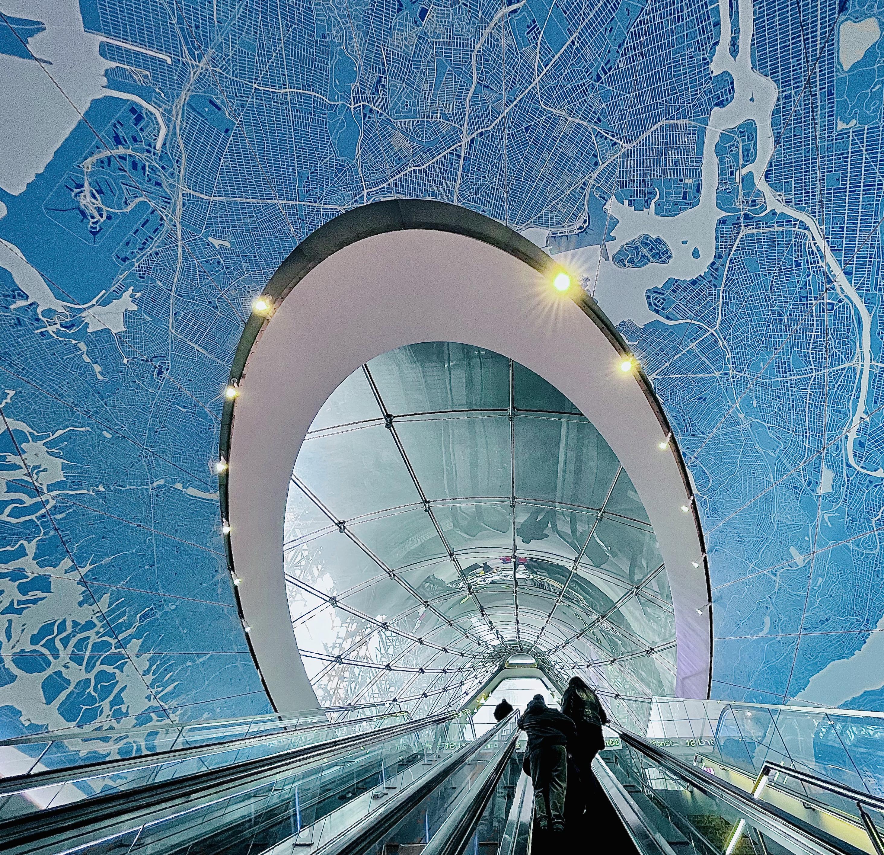 A color, wide-angle photograph in square aspect ratio that captures an upward-facing perspective from a moving escalator inside Moynihan Train Hall in Manhattan. Two commuters are seen from behind, riding the escalator toward a brightly lit exit, their silhouettes framed by the expansive architectural elements surrounding them. The escalator stretches upward in a symmetrical V-shape, flanked by gleaming metallic handrails and glass panels that reflect the ambient lighting. Dominating the upper portion of the frame is a striking blue cartographic art installation that sprawls across the ceiling and walls, depicting a detailed street grid and waterways resembling a map of New York City. Overlaying this vibrant blue map is a massive, curved glass architectural archway with warm, circular recessed lights that trace its perimeter.