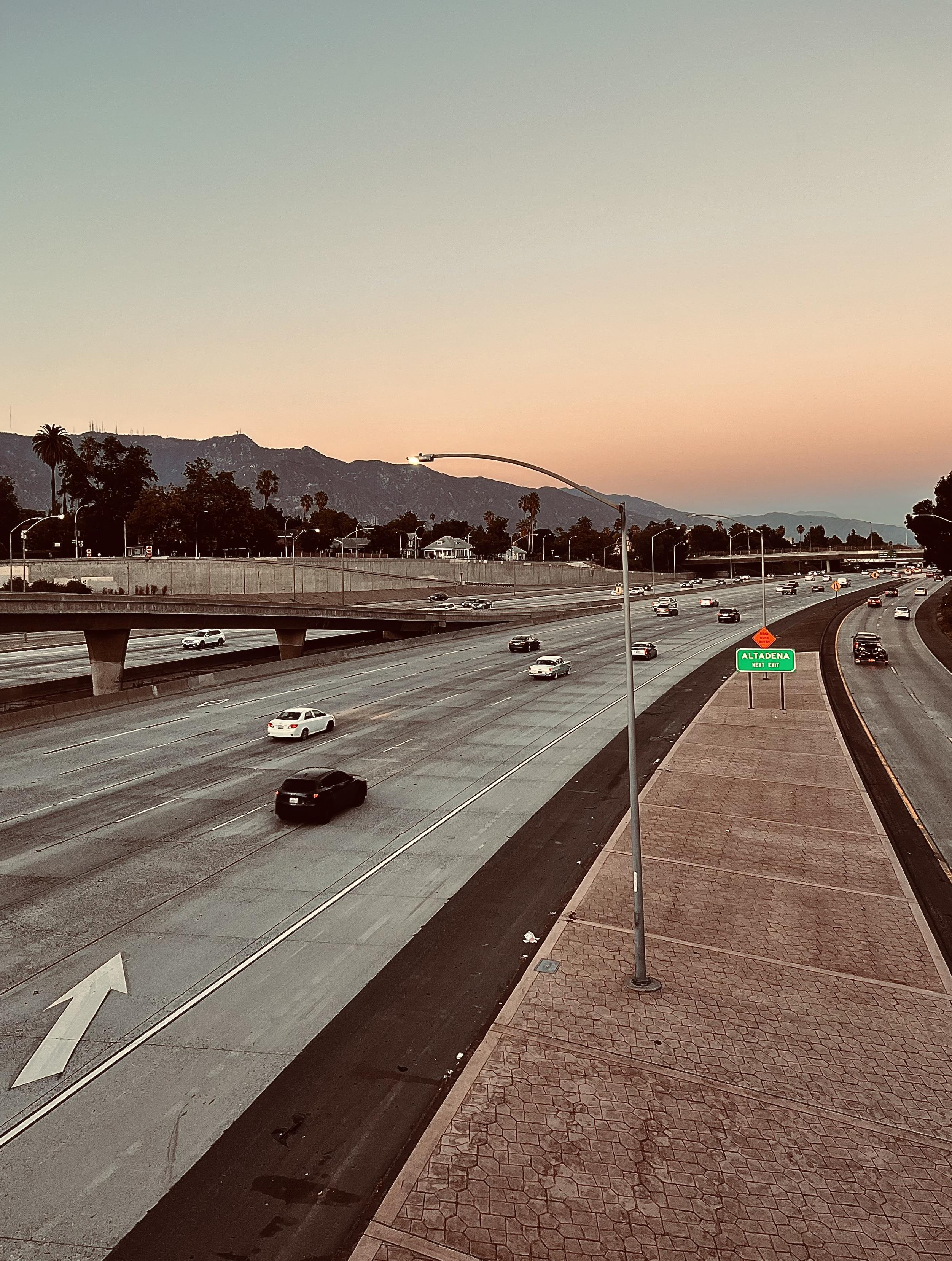 A color, wide-angle photograph in portrait orientation that captures a wide, multi-lane highway curving slightly to the right under a soft, gradient sunset sky. The upper section of the frame is dominated by a peaceful twilight sky that transitions from a pale peach near the horizon to a cool lavender gray higher up. Silhouetted against this colorful backdrop are the distant, rugged peaks of mountains stretching horizontally across the background, dotted with scattered palm trees and sparse vegetation along their base. An overpass bridge spans across the left side of the multi-lane freeway, with various cars traveling along both the main road and the elevated ramp. In the foreground, a paved concrete shoulder runs parallel to the highway, featuring a curved metal light pole standing tall near the center and a green highway sign reading "Altadena" just ahead. The concrete lanes of the freeway are marked with crisp white lane lines and a large white directional arrow painted on the far-left lane. Several vehicles, including a prominent dark SUV and a white sedan, drive along the lanes, casting soft shadows as the evening light begins to dim. The entire scene is bathed in a warm, muted color palette.