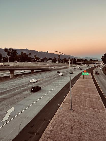 A color, wide-angle photograph in portrait orientation that captures a wide, multi-lane highway curving slightly to the right under a soft, gradient sunset sky. The upper section of the frame is dominated by a peaceful twilight sky that transitions from a pale peach near the horizon to a cool lavender gray higher up. Silhouetted against this colorful backdrop are the distant, rugged peaks of mountains stretching horizontally across the background, dotted with scattered palm trees and sparse vegetation along their base. An overpass bridge spans across the left side of the multi-lane freeway, with various cars traveling along both the main road and the elevated ramp. In the foreground, a paved concrete shoulder runs parallel to the highway, featuring a curved metal light pole standing tall near the center and a green highway sign reading "Altadena" just ahead. The concrete lanes of the freeway are marked with crisp white lane lines and a large white directional arrow painted on the far-left lane. Several vehicles, including a prominent dark SUV and a white sedan, drive along the lanes, casting soft shadows as the evening light begins to dim. The entire scene is bathed in a warm, muted color palette.