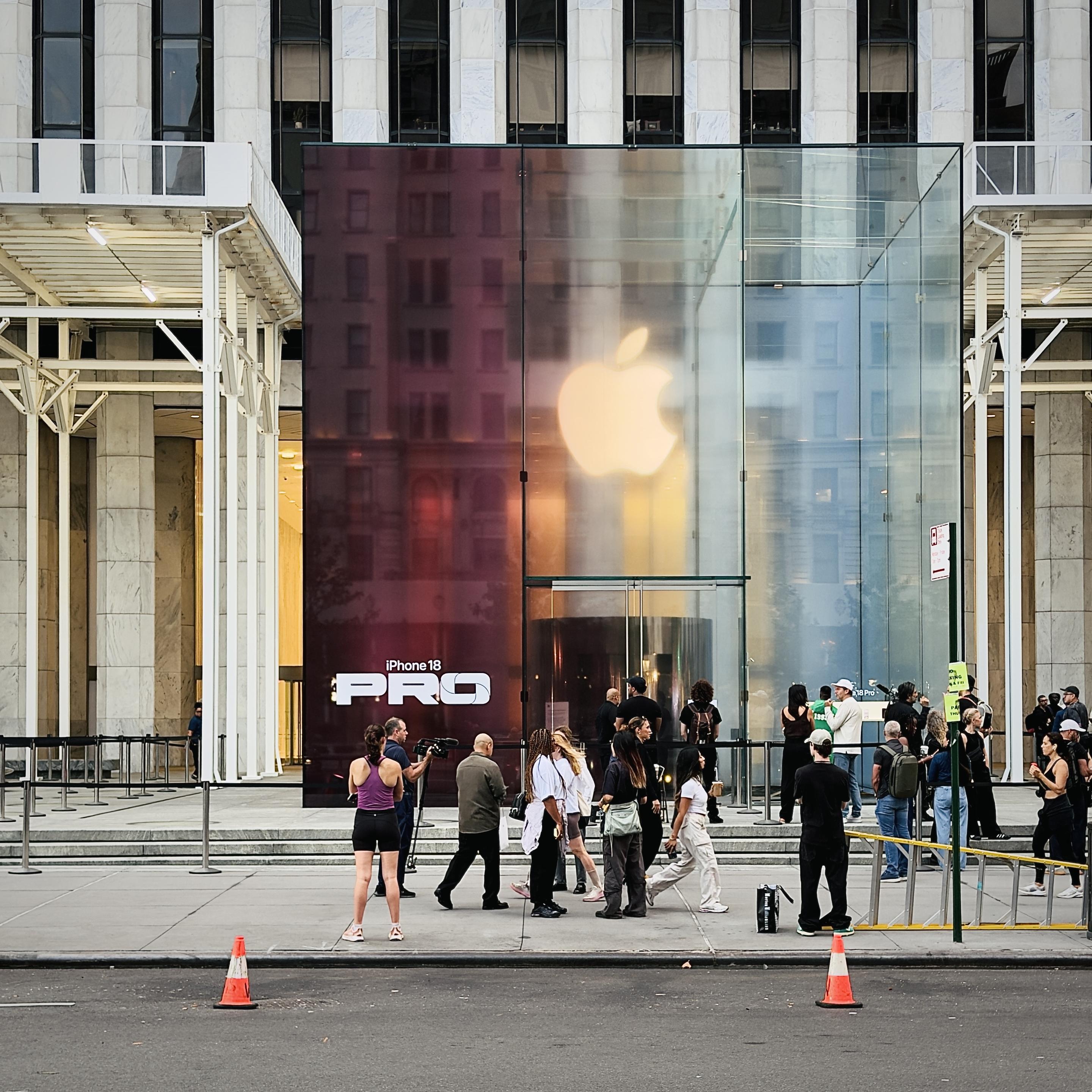 A color, wide-angle photograph that captures the Fifth Avenue Apple Store glass cube in Manhattan, transformed for the iPhone 18 Pro launch. The structure has received a special multi-toned color makeover, featuring a deep burgundy on the left panel emblazoned with a prominent white Apple logo and the bold white text "iPhone 18 PRO" near the bottom. The adjacent glass panels transition into a warm golden hue and a clear, reflective glass finish on the right, revealing the glowing white Apple logo suspended overhead inside the entrance. In the foreground, a plaza stretches across the scene with a light gray stone sidewalk where a crowd of customers, media personnel, and staff members have gathered. Several people stand near metal stanchions and queue barriers, with a few individuals holding cameras or recording equipment. The background showcases the classical, columned stone architecture of surrounding Manhattan buildings under a bright sky, while two orange traffic cones sit on the dark asphalt street at the bottom edge of the frame.