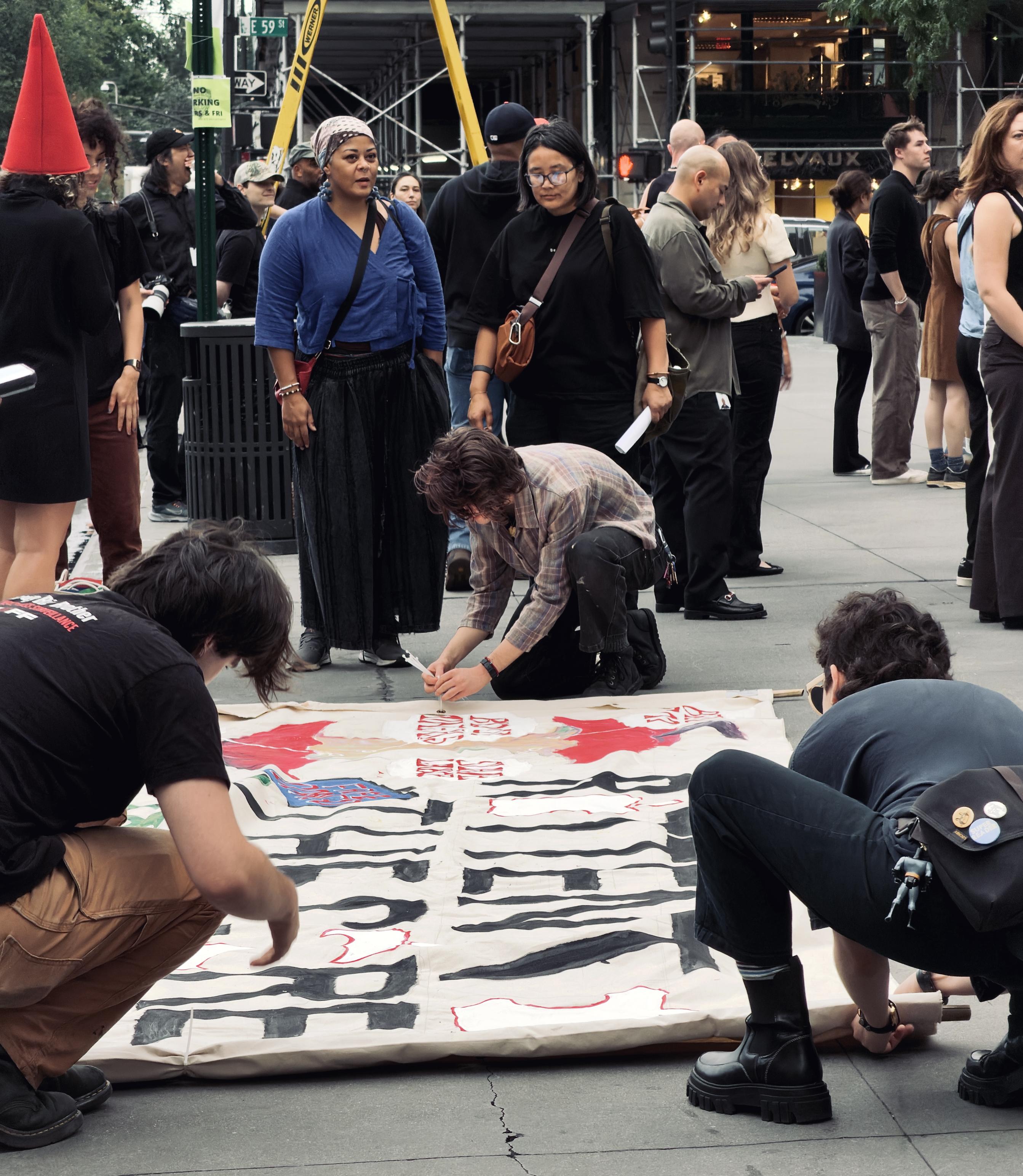 A color, wide-angle photograph in portrait orientation that captures several people gathered on a gray stone plaza, with three individuals kneeling on the ground in the foreground to paint a large white protest banner. The banner, stretched out horizontally on the pavement, features large, bold, block letters painted in black that read "ROTTEN TO THE CORE", accompanied by red painted motifs and smaller text. The person on the left wears a black t-shirt and tan pants, leaning forward to work on the edge of the fabric. In the center, a person in a plaid shirt is crouched down holding a paintbrush, while another person on the right, dressed in a dark top and jeans with platform boots, leans over the opposite side of the banner. Standing behind the banner is a diverse group of onlookers and participants watching the painting process unfold. Prominently in the middle-ground stand two women, one wearing a vibrant blue wrap top and dark patterned skirt, and beside her, a woman in a black shirt and glasses. Other pedestrians and bystanders are scattered in the background, some holding bags or looking on casually. The urban setting features architectural elements, scaffolding, and street signs overhead.