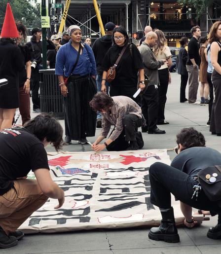 A color, wide-angle photograph in portrait orientation that captures several people gathered on a gray stone plaza, with three individuals kneeling on the ground in the foreground to paint a large white protest banner. The banner, stretched out horizontally on the pavement, features large, bold, block letters painted in black that read "ROTTEN TO THE CORE", accompanied by red painted motifs and smaller text. The person on the left wears a black t-shirt and tan pants, leaning forward to work on the edge of the fabric. In the center, a person in a plaid shirt is crouched down holding a paintbrush, while another person on the right, dressed in a dark top and jeans with platform boots, leans over the opposite side of the banner. Standing behind the banner is a diverse group of onlookers and participants watching the painting process unfold. Prominently in the middle-ground stand two women, one wearing a vibrant blue wrap top and dark patterned skirt, and beside her, a woman in a black shirt and glasses. Other pedestrians and bystanders are scattered in the background, some holding bags or looking on casually. The urban setting features architectural elements, scaffolding, and street signs overhead.