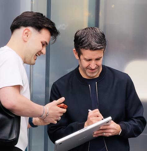 A color, medium-angle photograph in square aspect ratio that captures John Ternus, Apple's CEO, standing outside the Fifth Avenue Apple Store during the iPhone 18 Pro launch event. Ternus, with neatly styled dark-graying hair and a thin stubble, wears a dark zippered bomber jacket over a navy shirt. He is focused intently downward, holding a sharpie in his right hand as he palms a silver MacBook Pro in his left hand. Standing to his left is a young man with a short, tapered haircut, wearing a white crew-neck t-shirt and a dark shoulder bag. The customer looks on with an enthusiastic, smiling expression, holding an orange iPhone in one hand while watching Ternus sign his laptop. The background features the reflective glass panels and muted structural elements of the store.