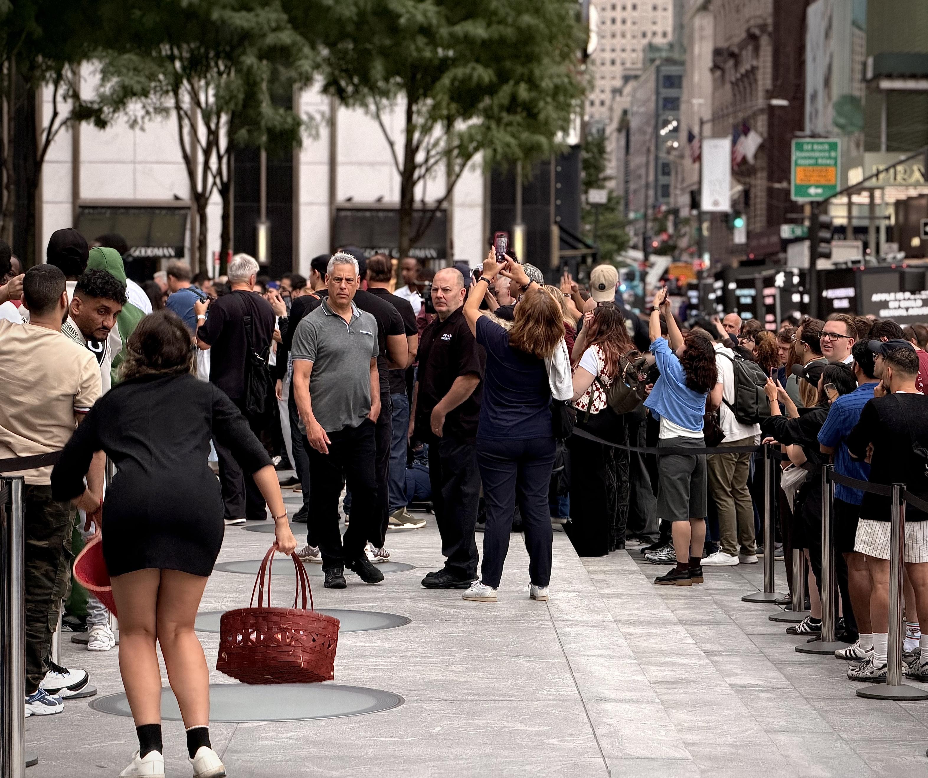 A color, wide-angle photograph in landscape orientation that captures a bustling plaza and sidewalk outside the flagship Fifth Avenue Apple Store during the iPhone 18 Pro product launch, where a protester is being noticed prior to being intercepted by security personnel. In the foreground, a woman is facing away from the camera dressed in a short black dress and white sneakers, caught mid-stride while holding a large, woven reddish basket. Several security staff members and onlookers are tracking her movement, with a security guard in a dark polo shirt and black pants standing alertly near the center of the frame. The textured stone pavement of the plaza stretches across the lower half of the image, marked by circular architectural inlays, while metal stanchions and black ropes manage the dense crowd. The background reveals a dense urban streetscape filled with a large queue of customers and spectators, many of whom are holding up smartphones to capture the scene. Tall commercial buildings, storefronts, and distant street signs rise vertically under a bright, overcast daylight sky.