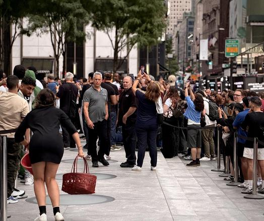 A color, wide-angle photograph in landscape orientation that captures a bustling plaza and sidewalk outside the flagship Fifth Avenue Apple Store during the iPhone 18 Pro product launch, where a protester is being noticed prior to being intercepted by security personnel. In the foreground, a woman is facing away from the camera dressed in a short black dress and white sneakers, caught mid-stride while holding a large, woven reddish basket. Several security staff members and onlookers are tracking her movement, with a security guard in a dark polo shirt and black pants standing alertly near the center of the frame. The textured stone pavement of the plaza stretches across the lower half of the image, marked by circular architectural inlays, while metal stanchions and black ropes manage the dense crowd. The background reveals a dense urban streetscape filled with a large queue of customers and spectators, many of whom are holding up smartphones to capture the scene. Tall commercial buildings, storefronts, and distant street signs rise vertically under a bright, overcast daylight sky.