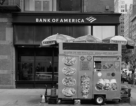 A noir, wide-angle photograph in landscape orientation that captures a Halal food cart stationed on a Manhattan sidewalk directly in front of a Bank of America branch. The food cart is the prominent central focus, featuring three striped Sabrett umbrellas overhead and a menu display showcasing pictures of rice platters, gyros, and burgers alongside a vertical image of a vertical spit of meat. Signage on the cart details food options and a phone number, while a small traffic cone sits near its front on the concrete pavement. The background is dominated by the storefront of the bank, with the prominent "BANK OF AMERICA" lettering and corporate logo standing out against the fascia. Reflections are visible in the large glass windows behind the cart, and the narrow urban street extends into the distance on the right, where pedestrians walk past parked vehicles.