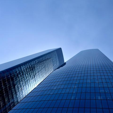 A color, wide-angle photograph in square aspect ratio, a low-angle view that captures two massive glass skyscrapers soaring upward against a clear, pale blue sky. The structures are sleek, dark blue curtain walls made of reflective glass panels and gridded window frames, and dominate the composition. The skyscraper on the right features a smooth, gently curved facade that catches the bright daylight, reflecting a uniform blue tone across its grid. In contrast, the building on the left exhibits a more angled, textured surface, with strong light reflections and shadows playing across its surface, creating a dynamic visual interplay between the two.