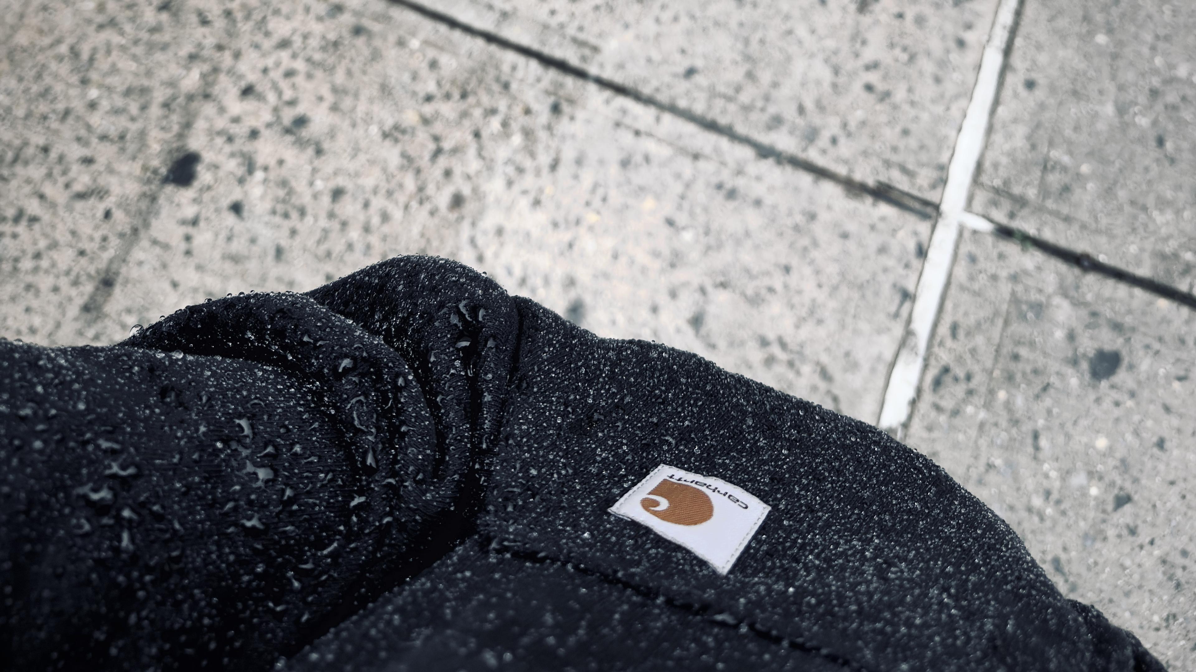 A color, medium-angle, close-up photograph in landscape orientation that captures a dark-colored Carhartt hoodie drenched in a heavy downpour. The textured, water-repellent fabric of the sweatshirt is densely speckled with countless glistening water droplets and larger beads of moisture clinging. A rectangular white Carhartt fabric patch featuring the brand's iconic brown "C" logo and script lettering is stitched onto the upper left pocket area. The shot is framed from a first-person perspective looking down at my lower chest and left sleeve. In the background, the wet, textured concrete sidewalk stretches out diagonally across the upper portion of the frame.