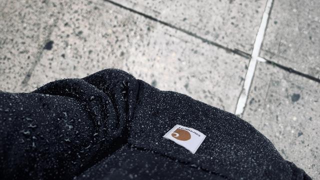 A color, medium-angle, close-up photograph in landscape orientation that captures a dark-colored Carhartt hoodie drenched in a heavy downpour. The textured, water-repellent fabric of the sweatshirt is densely speckled with countless glistening water droplets and larger beads of moisture clinging. A rectangular white Carhartt fabric patch featuring the brand's iconic brown "C" logo and script lettering is stitched onto the upper left pocket area. The shot is framed from a first-person perspective looking down at my lower chest and left sleeve. In the background, the wet, textured concrete sidewalk stretches out diagonally across the upper portion of the frame.