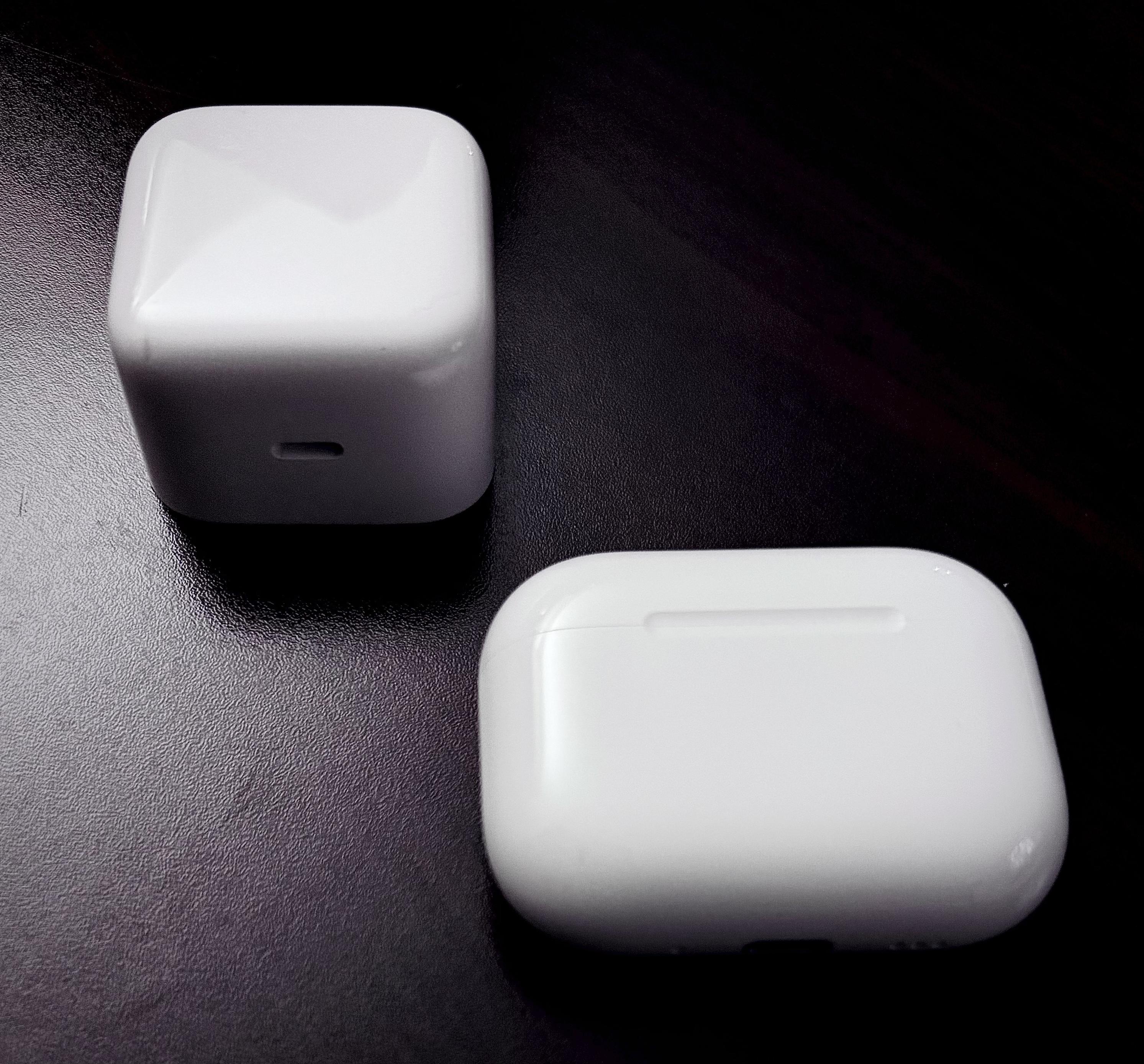 A color, medium-angle photograph in square aspect ratio. A desaturated scene of a white power adapter and AirPods case, both USB-C, sitting on a laminate table under direct and moody lighting.