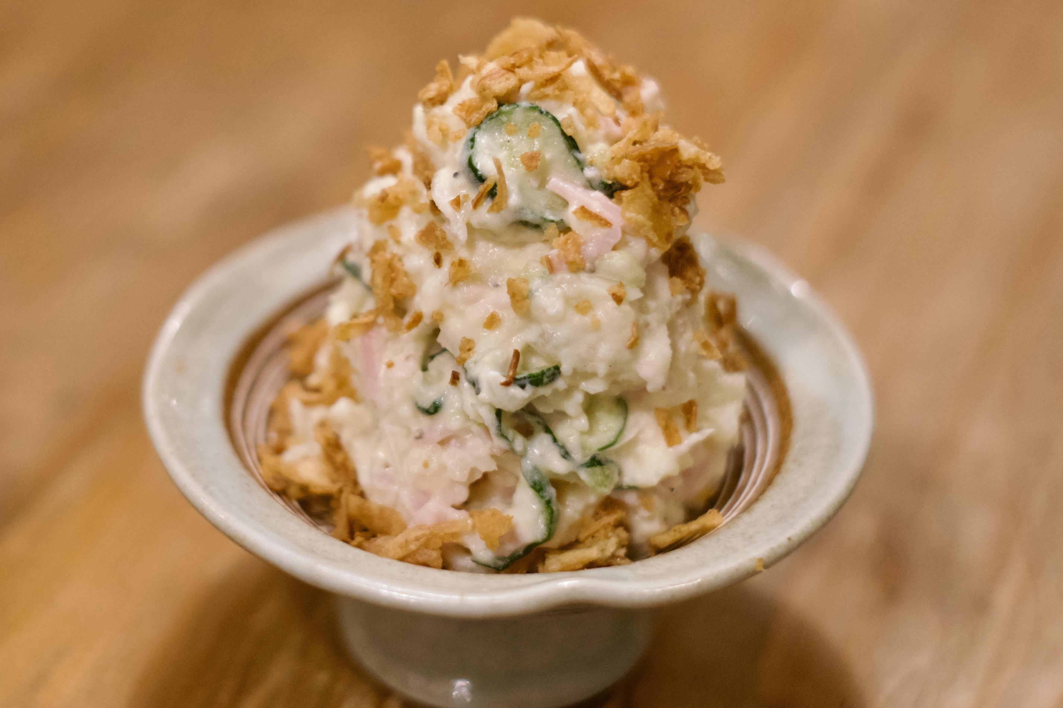 A color, medium-angle, close-up photograph in landscape orientation that captures a neat, tall mound of Japanese-style potato salad served in a light-colored, shallow ceramic pedestal bowl. The potato salad has a creamy, textured appearance, with thin slices of vibrant green cucumber and pink onion visibly folded throughout. The entire mound is generously garnished with a scattering of golden-brown, crispy onion flakes that add a contrasting texture and rustic appeal. The bowl rests on a warm-toned wooden surface, and the background is softly blurred to keep the focus entirely on the presentation.