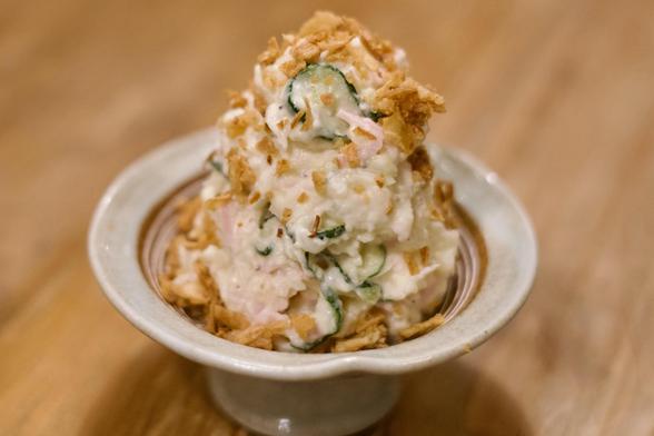 A color, medium-angle, close-up photograph in landscape orientation that captures a neat, tall mound of Japanese-style potato salad served in a light-colored, shallow ceramic pedestal bowl. The potato salad has a creamy, textured appearance, with thin slices of vibrant green cucumber and pink onion visibly folded throughout. The entire mound is generously garnished with a scattering of golden-brown, crispy onion flakes that add a contrasting texture and rustic appeal. The bowl rests on a warm-toned wooden surface, and the background is softly blurred to keep the focus entirely on the presentation.