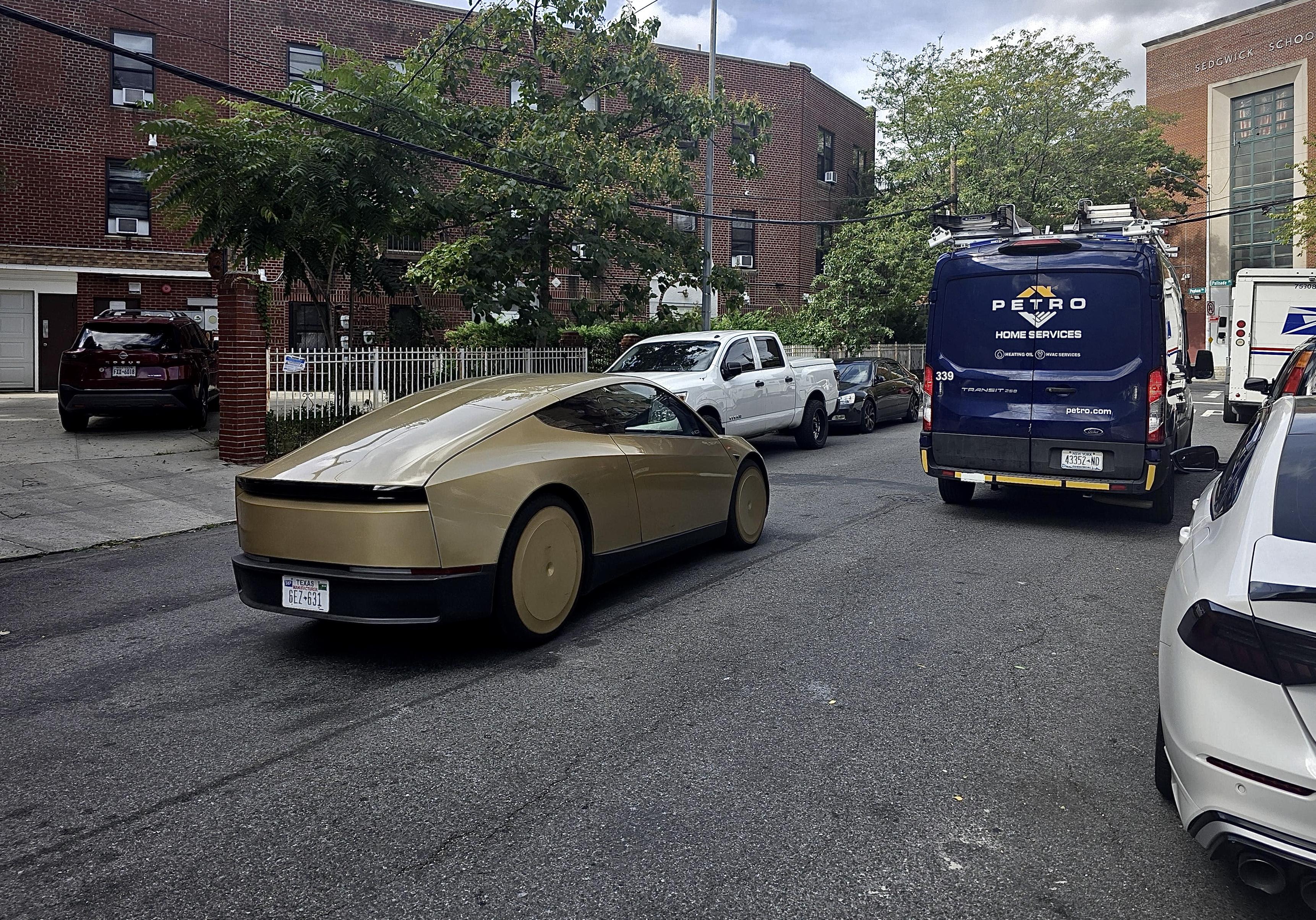 A color, wide-angle photograph in landscape orientation that captures a metallic gold Tesla Cybercabdriving down a residential Bronx street on a clear day. The vehicle features an aerodynamic coupe-like profile with a heavily sloped roofline, a smooth rear light bar, and gold-covered wheels. It is positioned diagonally in the center-left of the frame, angled away from the viewer. A Texas rear license plate is visible. The surrounding environment consists of a typical New York City neighborhood street lined with parked vehicles, including a white pickup truck, a dark sedan, a blue "Petro Home Services" utility van on the right, and a white postal delivery truck further ahead. In the background, traditional multi-story red brick apartment buildings and institutional brick architecture are partially obscured by green leafy trees and overhead power lines. The foreground features a small portion of a white car's bumper and hood on the lower right, while the asphalt road displays a slightly grainy texture with tire marks.