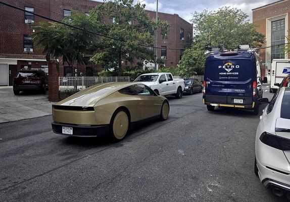 A color, wide-angle photograph in landscape orientation that captures a metallic gold Tesla Cybercabdriving down a residential Bronx street on a clear day. The vehicle features an aerodynamic coupe-like profile with a heavily sloped roofline, a smooth rear light bar, and gold-covered wheels. It is positioned diagonally in the center-left of the frame, angled away from the viewer. A Texas rear license plate is visible. The surrounding environment consists of a typical New York City neighborhood street lined with parked vehicles, including a white pickup truck, a dark sedan, a blue "Petro Home Services" utility van on the right, and a white postal delivery truck further ahead. In the background, traditional multi-story red brick apartment buildings and institutional brick architecture are partially obscured by green leafy trees and overhead power lines. The foreground features a small portion of a white car's bumper and hood on the lower right, while the asphalt road displays a slightly grainy texture with tire marks.