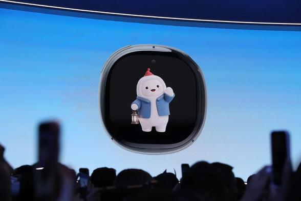 A color, wide-angle photograph that captures a press audience holding up phones and cameras to take photos of a keynote screen displaying a Meta Muse AI dedicated hardware gadget. The avatar on screen looks like a weird snow man wearing a denim jacket and a tiny red hat and carrying a lantern.