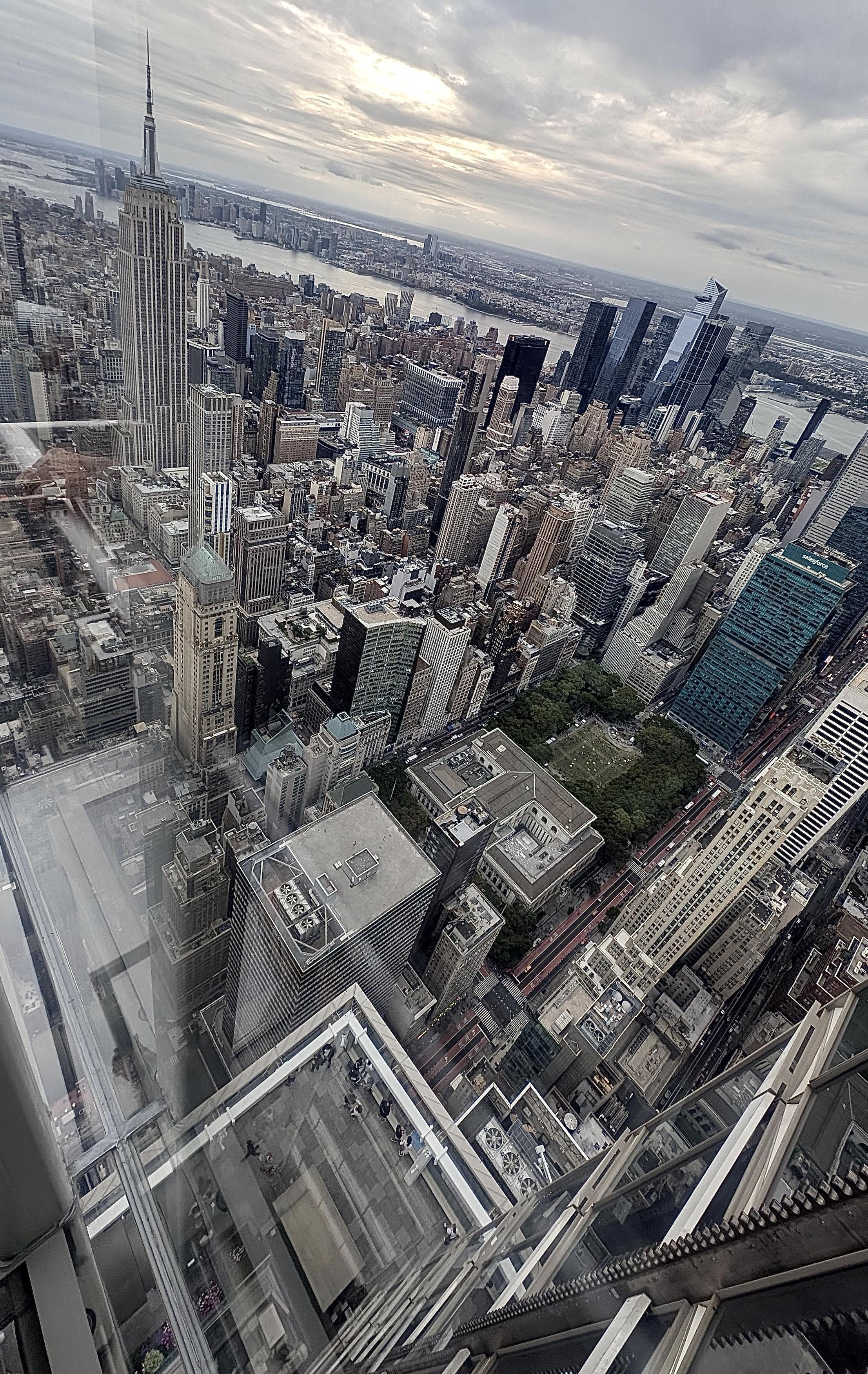 A color, wide-angle photograph in portrait orientation the captures an elevated, high-angle view from an observation deck overlooking Manhattan. The Empire State Building stands prominently on the mid-left side, its distinct spire pointing upward under an overcast sky. The surrounding cityscape is a dense grid of towering skyscrapers, commercial buildings, and smaller structures stretching out toward the Hudson river in the distance. A patch of green park space at Bryant Park is visible amidst the concrete, while modern glass high-rises, including the Salesforce Tower, rise toward the right. The foreground reveals the structural framing of the observation deck itself, with clear glass panels, metal railings, and a direct view looking straight down toward the street level far below. The lighting is diffused and soft, characteristic of an overcast day, casting subtle shadows across the tightly packed architecture.