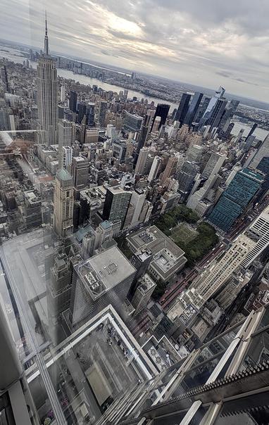 A color, wide-angle photograph in portrait orientation the captures an elevated, high-angle view from an observation deck overlooking Manhattan. The Empire State Building stands prominently on the mid-left side, its distinct spire pointing upward under an overcast sky. The surrounding cityscape is a dense grid of towering skyscrapers, commercial buildings, and smaller structures stretching out toward the Hudson river in the distance. A patch of green park space at Bryant Park is visible amidst the concrete, while modern glass high-rises, including the Salesforce Tower, rise toward the right. The foreground reveals the structural framing of the observation deck itself, with clear glass panels, metal railings, and a direct view looking straight down toward the street level far below. The lighting is diffused and soft, characteristic of an overcast day, casting subtle shadows across the tightly packed architecture.