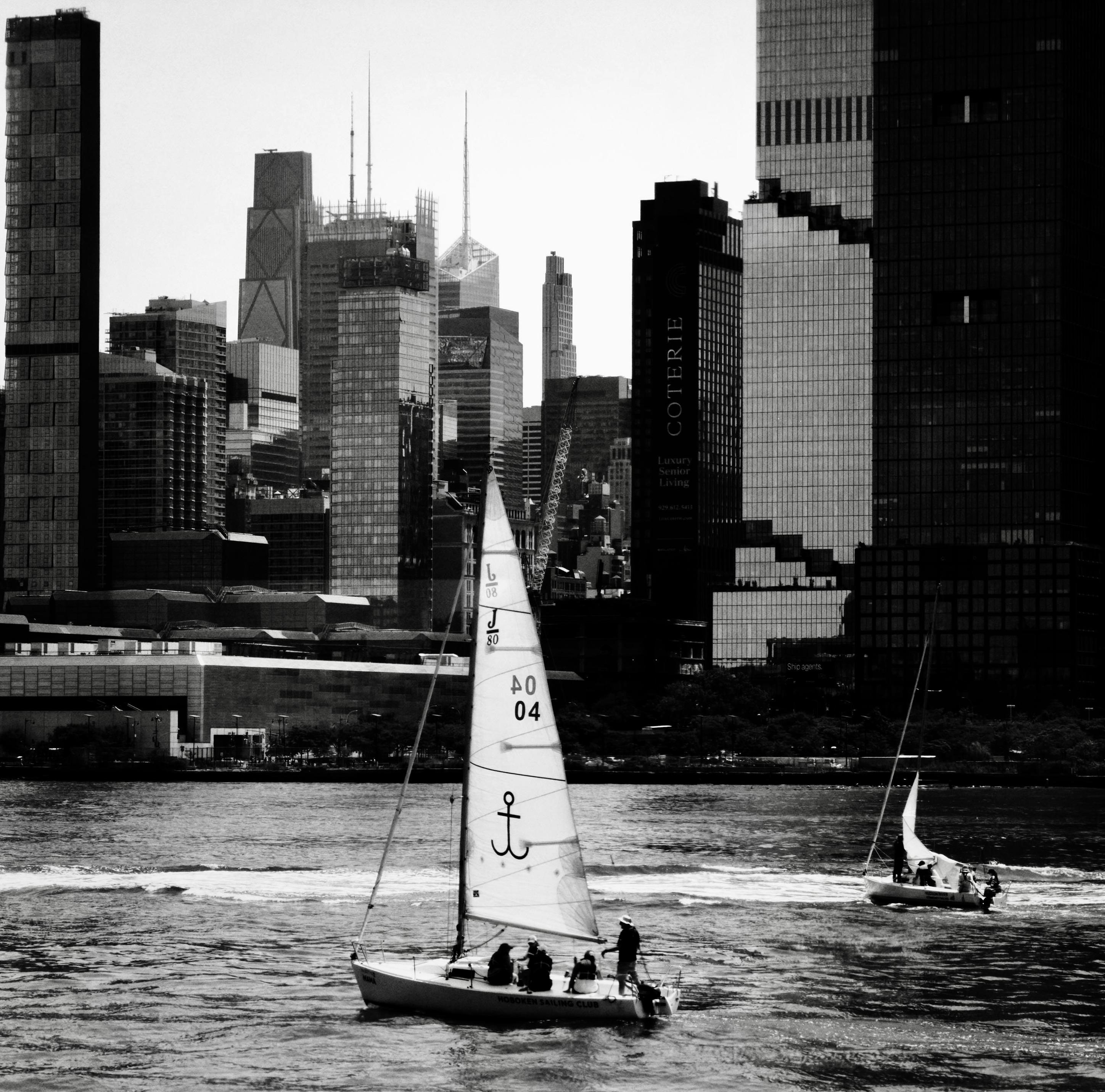 A noir, wide-angle photograph in square aspect ratio that captures a morning scene along the Hudson River with two sailboats navigating the choppy waters. In the foreground, a prominent sailboat moves across the lower-center portion of the frame, its large, white sail featuring distinctive markings including an anchor symbol, numerical digits, and letters. Several people are visible on the deck of this primary boat, with one person standing near the stern holding the steering column. To the right and slightly further back, a smaller sailboat with its white sail unfurled tilts gently as it cuts through the water. The background is dominated by the dense, towering skyscrapers of the Manhattan skyline under a bright, overcast sky. The architecture showcases a dramatic variety of high-rise building styles and heights, ranging from sleek, modern glass towers to older, structured facades. The water in the river shows visible ripples and waves, catching the ambient light while the entire composition is rendered in high-contrast monochrome tones.
