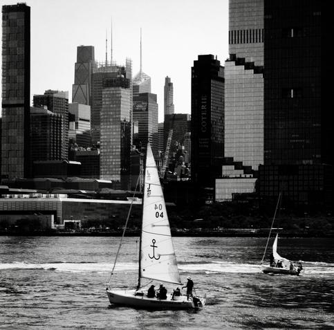 A noir, wide-angle photograph in square aspect ratio that captures a morning scene along the Hudson River with two sailboats navigating the choppy waters. In the foreground, a prominent sailboat moves across the lower-center portion of the frame, its large, white sail featuring distinctive markings including an anchor symbol, numerical digits, and letters. Several people are visible on the deck of this primary boat, with one person standing near the stern holding the steering column. To the right and slightly further back, a smaller sailboat with its white sail unfurled tilts gently as it cuts through the water. The background is dominated by the dense, towering skyscrapers of the Manhattan skyline under a bright, overcast sky. The architecture showcases a dramatic variety of high-rise building styles and heights, ranging from sleek, modern glass towers to older, structured facades. The water in the river shows visible ripples and waves, catching the ambient light while the entire composition is rendered in high-contrast monochrome tones.