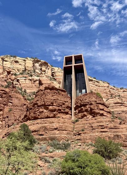 A color, wide-angle photograph in portrait orientation that captures the Chapel of the Holy Cross in Sedona, Arizona from a low-angle perspective. The chapel is built directly into the striking red rock buttes of the desert landscape. The building features a minimalist, modern architectural style with a prominent concrete exterior framing a massive, dark-tinted glass cross that forms its front facade. The structure appears tightly nestled between two towering red rock formations that match its earthy, rust-colored hues. The surrounding landscape includes sparse desert vegetation, such as green shrubs, low bushes, and cacti scattered across the reddish-brown terrain in the foreground. Above the geological formations, a vibrant blue sky fills the upper portion of the frame, lightly scattered with wispy white clouds. The bright daylight casts gentle shadows that emphasize the layered textures and rugged crevices of the sandstone cliffs.