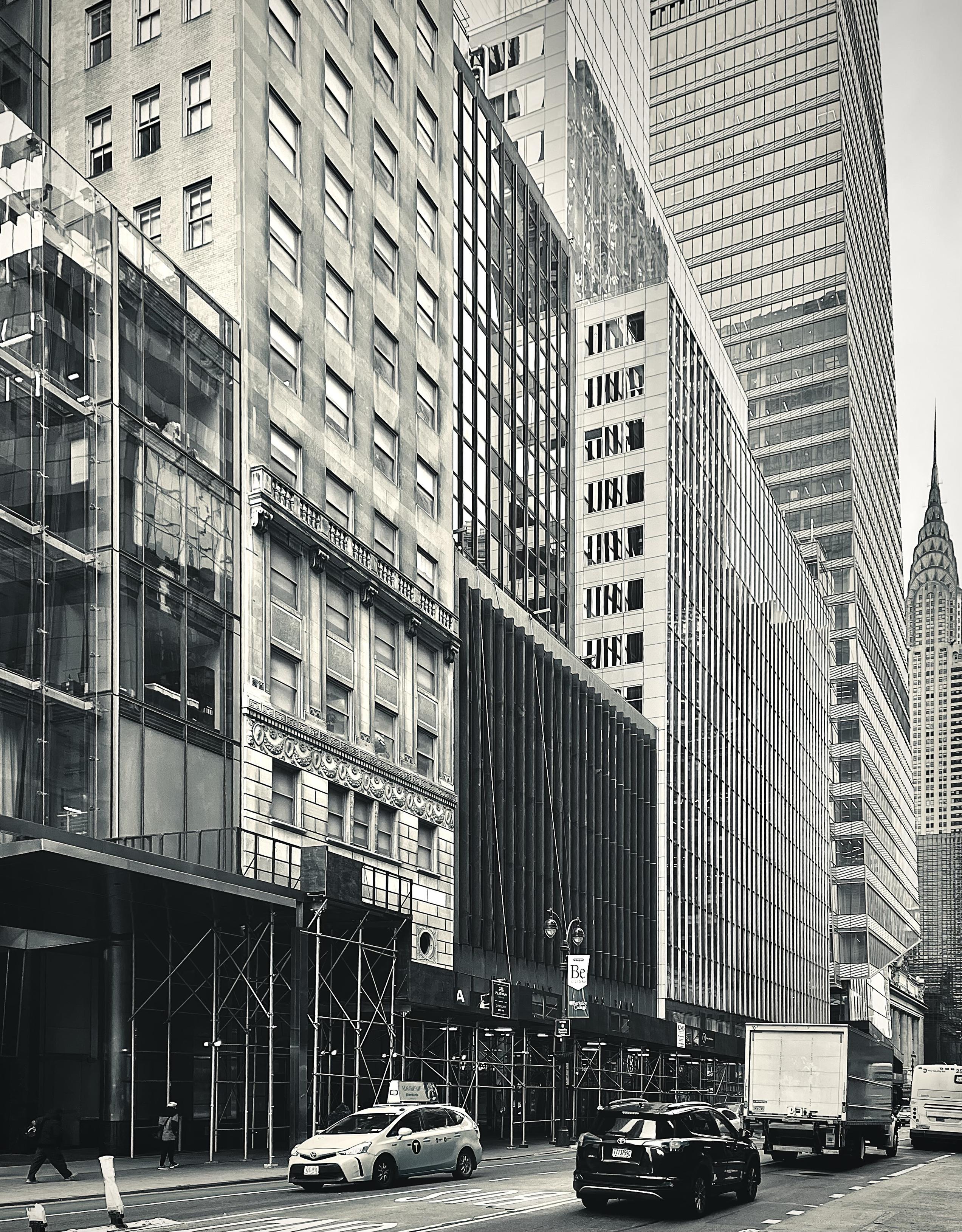 A noir, wide-angle photograph in portrait orientation that captures a view looking down 42nd Street toward the Chrysler Building in Midtown Manhattan. The composition is dominated on the left and center by a dense architectural wall of contrasting buildings showcasing diverse facades, ranging from modern glass-front structures and classic stone masonry with intricate ornamentation to vertical louvered panels and contemporary high-rise towers. Scaffolding lines the base of several buildings on the lower left, where pedestrians walk along the sidewalk next to the curb. The street level features light and dark passenger cars, including a taxi, moving alongside a large delivery truck heading down the multi-lane roadway. Farther down the corridor on the right side, towering skyscrapers recede into the distance, culminating in the distinct terraced spire and crown of the Chrysler Building, which stands out prominently against the pale sky.
