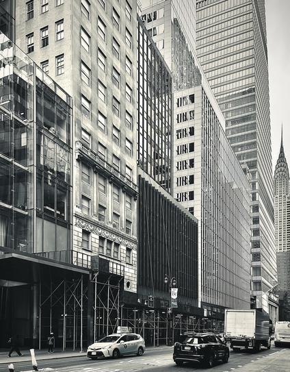 A noir, wide-angle photograph in portrait orientation that captures a view looking down 42nd Street toward the Chrysler Building in Midtown Manhattan. The composition is dominated on the left and center by a dense architectural wall of contrasting buildings showcasing diverse facades, ranging from modern glass-front structures and classic stone masonry with intricate ornamentation to vertical louvered panels and contemporary high-rise towers. Scaffolding lines the base of several buildings on the lower left, where pedestrians walk along the sidewalk next to the curb. The street level features light and dark passenger cars, including a taxi, moving alongside a large delivery truck heading down the multi-lane roadway. Farther down the corridor on the right side, towering skyscrapers recede into the distance, culminating in the distinct terraced spire and crown of the Chrysler Building, which stands out prominently against the pale sky.