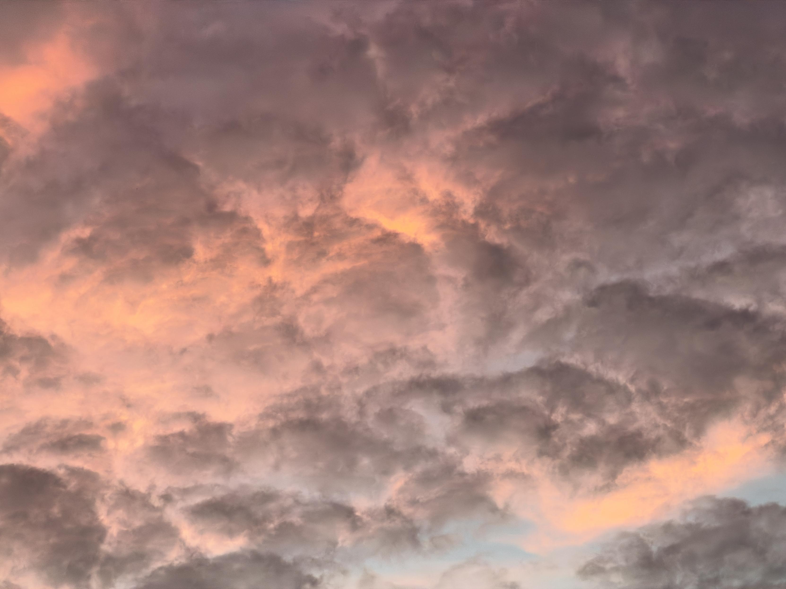 A color, wide-angle photograph in landscape orientation that captures a dense, dramatic cloudscape filled with moody, textured clouds stretching across the Manhattan sky. The lower and upper portions of the sky are heavily layered with charcoal-gray and muted purple clouds that roll and overlap in a dense formation, creating a soft yet heavy appearance.nPiercing through the dark cloud cover are warm patches of golden-orange and soft pink light, highlighting the edges and inner textures of the cloud formations. The interplay between the dark, shadowed masses and the vibrant breaks gives the entire sky a luminous and dynamic quality.