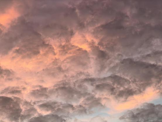 A color, wide-angle photograph in landscape orientation that captures a dense, dramatic cloudscape filled with moody, textured clouds stretching across the Manhattan sky. The lower and upper portions of the sky are heavily layered with charcoal-gray and muted purple clouds that roll and overlap in a dense formation, creating a soft yet heavy appearance.nPiercing through the dark cloud cover are warm patches of golden-orange and soft pink light, highlighting the edges and inner textures of the cloud formations. The interplay between the dark, shadowed masses and the vibrant breaks gives the entire sky a luminous and dynamic quality.
