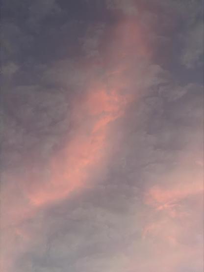 A color, wide-angle photograph in portrait orientation that captures a dramatic sky filled with thick, textured clouds illuminated by sunset over Manhattan. The dominant feature is a diagonal band of vibrant, warm pink and peach light cutting through the center of the frame, contrasting against cooler, muted grayish-purple and dark blue clouds that fill the rest of the sky.