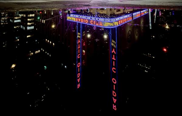 A color, wide-angle photograph taken at night in New York City that captures the facade and neon signage of Radio City Music Hall inverted in a puddle on the street, framed by a narrow strip of dark pavement at the very top. The water's surface creates a mirror-like reflection where the Art Deco building appears upside down. Dominating the center of the reflection are two vertical neon signs glowing in bright red, spelling out "RADIO CITY" from top to bottom, flanked by a sweeping, multi-tiered marquee wrapped in bright purple and blue lights. The background features deep shadows contrasting with scattered yellow lights from nearby city windows and streetlights.