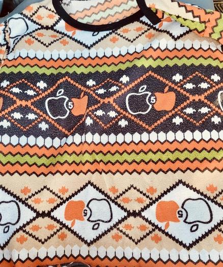 A color, medium-angle photograph in portrait orientation of an “ugly sweater” with a white, orange and green color scheme. Apple, Inc logos adorn the sweater in horizontal stripes. The number 5 appears next to the Apple logos, signifying the five year anniversary of the iMac.