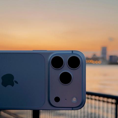 A color, wide-angle photograph in square aspect ratio that captures a scene along the Hudson River at sunrise. A glacier-colored iPhone 18 Pro is held horizontally in the foreground, dominating the lower left and center of the frame. The frosted, pale blue-grey back glass reflects the warm morning glow, featuring the Apple logo on the left and a raised, square camera module on the right housing three distinct lenses, a microphone hole, and a flash. The background captures a serene sunrise over the Hudson River, with a soft gradient sky shifting from warm orange near the horizon to pale blue above. Across the water, the hazy silhouette of a distant city skyline is visible, while a dark metal railing runs along the foreground waterfront, softly blurring into the glowing water and atmosphere.