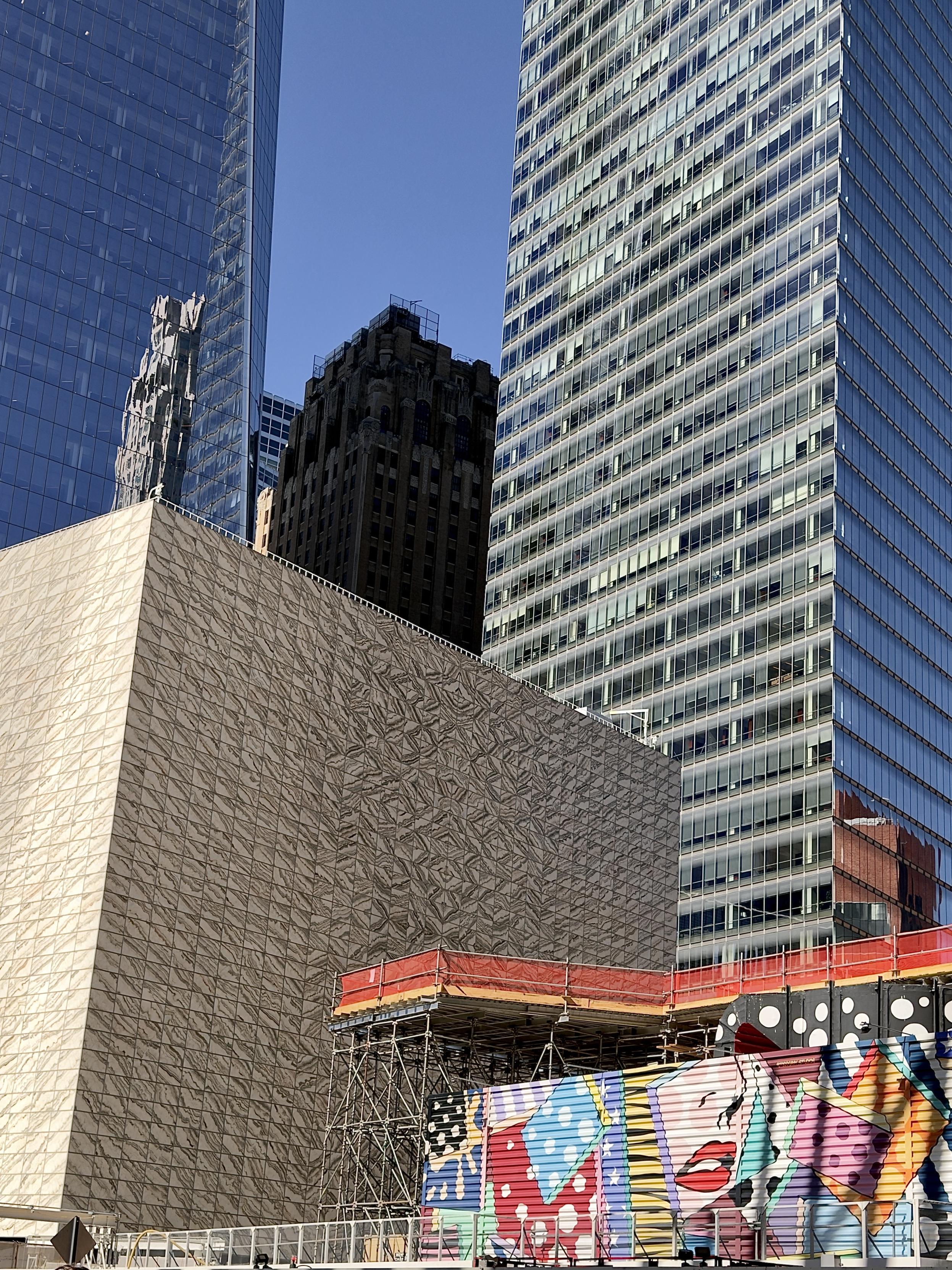 A color, wide-angle photograph in portrait orientation that captures a dynamic architectural composition outside the World Trade Center complex in New York City, set against a clear blue sky. Dominating the lower left portion of the frame is the distinctive cube-shaped building of the Perelman Performing Arts Center, featuring a striking facade of veined, translucent marble panels. Rising directly behind it in the center is a darker, classical Art Deco-style masonry skyscraper, which creates a sharp textural and historical contrast against the surrounding modern high-rises. Flanking the composition are two towering, contemporary glass skyscrapers with gridded curtain walls that reflect the daylight and surrounding structures. In the lower right foreground, a construction or staging area is visible, partially obscured by scaffolding and a vibrant, colorful mural featuring pop-art graphics, bold patterns, and a large rendering of red lips. The interplay of marble, weathered stone, sleek glass, and colorful street art highlights the diverse textures of the evolving downtown landscape.