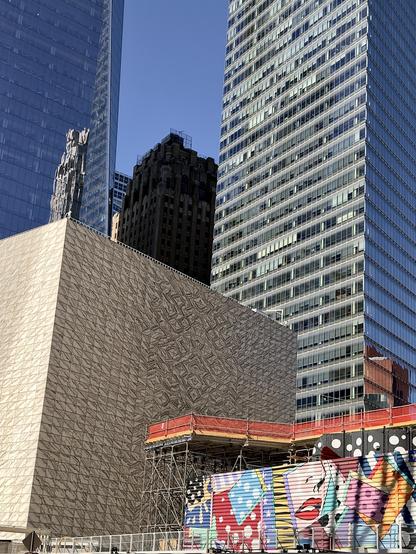 A color, wide-angle photograph in portrait orientation that captures a dynamic architectural composition outside the World Trade Center complex in New York City, set against a clear blue sky. Dominating the lower left portion of the frame is the distinctive cube-shaped building of the Perelman Performing Arts Center, featuring a striking facade of veined, translucent marble panels. Rising directly behind it in the center is a darker, classical Art Deco-style masonry skyscraper, which creates a sharp textural and historical contrast against the surrounding modern high-rises. Flanking the composition are two towering, contemporary glass skyscrapers with gridded curtain walls that reflect the daylight and surrounding structures. In the lower right foreground, a construction or staging area is visible, partially obscured by scaffolding and a vibrant, colorful mural featuring pop-art graphics, bold patterns, and a large rendering of red lips. The interplay of marble, weathered stone, sleek glass, and colorful street art highlights the diverse textures of the evolving downtown landscape.