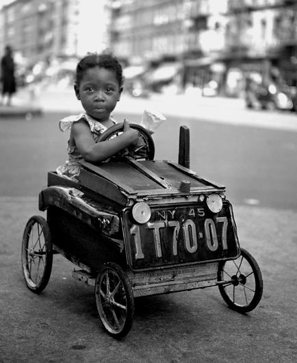 A noir, medium-angle photograph in square aspect ratio that captures a street scene in Harlem, Manhattan in 1947. A young Black girl sitting inside a handcrafted, box-shaped toy vehicle parked on a city street. She faces forward with a calm, wide-eyed expression, her hands resting around a circular steering wheel. She wears a patterned sleeveless summer dress with ruffled shoulders, and her dark hair is styled neatly. An ice cream cone is held in one hand. The wooden and metal toy car is detailed with repurposed components, featuring thin spoked wheels, a wooden crate base, a hinged hood structure, and a large black license plate attached to the front that reads "NY 45" above "1T70-07" flanked by two round headlights. The girl and her vehicle are centered in the frame on asphalt ground, with the surrounding neighborhood blurred soft-focus in the background. Multi-story residential brownstone and apartment buildings, storefront awnings, and traffic along the street line the background.