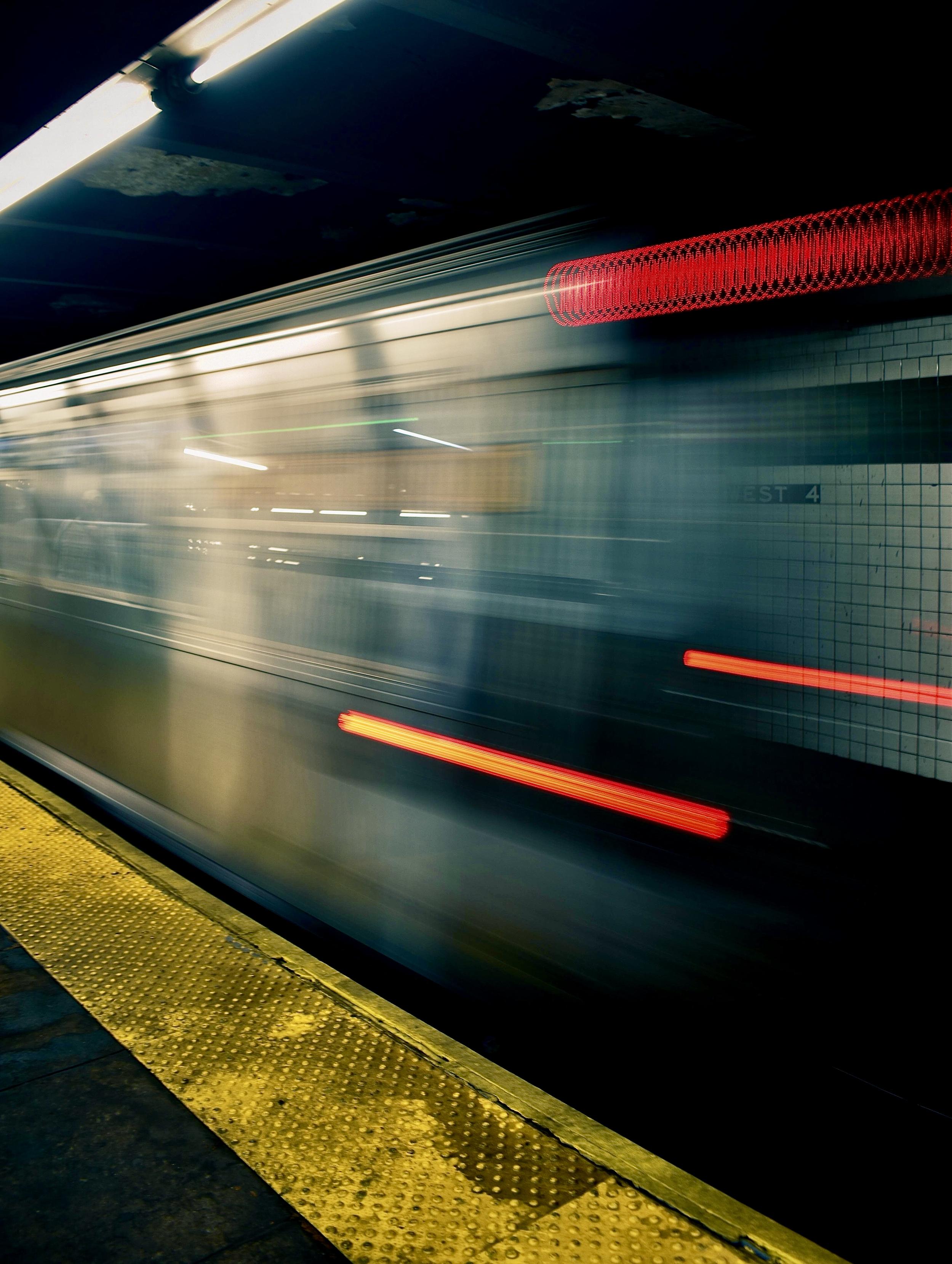 A color, wide-angle, long-exposure photograph that captures the high-speed motion of a train inside the West 4th Street subway station in Manhattan. In the bottom left foreground, a bright yellow textured tactile paving strip stretches diagonally along the edge of the platform. Overhead, a bright white fluorescent tube light casts a cool glow over the dark, aged ceiling and upper platform area. Across the tracks, white square subway tiles are partially visible through the translucency of the moving train, displaying the station name "WEST 4" marked on the wall.  The main subject of the image is the subway train, rendered as a dramatic, ghostly motion blur across the horizontal plane. Streak lights from the interior windows and bright red taillights trace glowing horizontal lines through the frame, conveying rapid speed and momentum. Near the upper right, an intricate coiled red light effect creates a spring-like pattern, while a vibrant red streak cuts horizontally across the middle right.