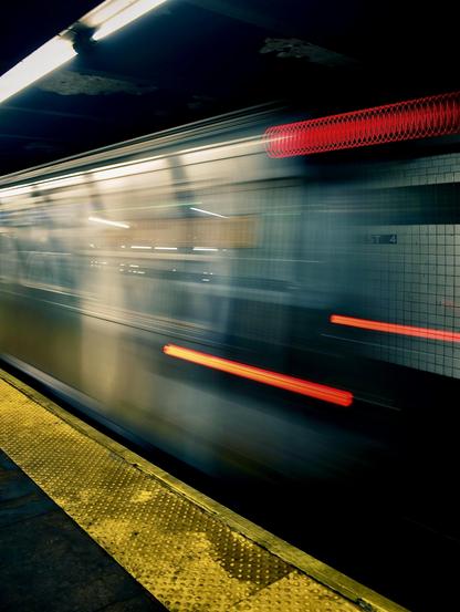 A color, wide-angle, long-exposure photograph that captures the high-speed motion of a train inside the West 4th Street subway station in Manhattan. In the bottom left foreground, a bright yellow textured tactile paving strip stretches diagonally along the edge of the platform. Overhead, a bright white fluorescent tube light casts a cool glow over the dark, aged ceiling and upper platform area. Across the tracks, white square subway tiles are partially visible through the translucency of the moving train, displaying the station name "WEST 4" marked on the wall.  The main subject of the image is the subway train, rendered as a dramatic, ghostly motion blur across the horizontal plane. Streak lights from the interior windows and bright red taillights trace glowing horizontal lines through the frame, conveying rapid speed and momentum. Near the upper right, an intricate coiled red light effect creates a spring-like pattern, while a vibrant red streak cuts horizontally across the middle right.