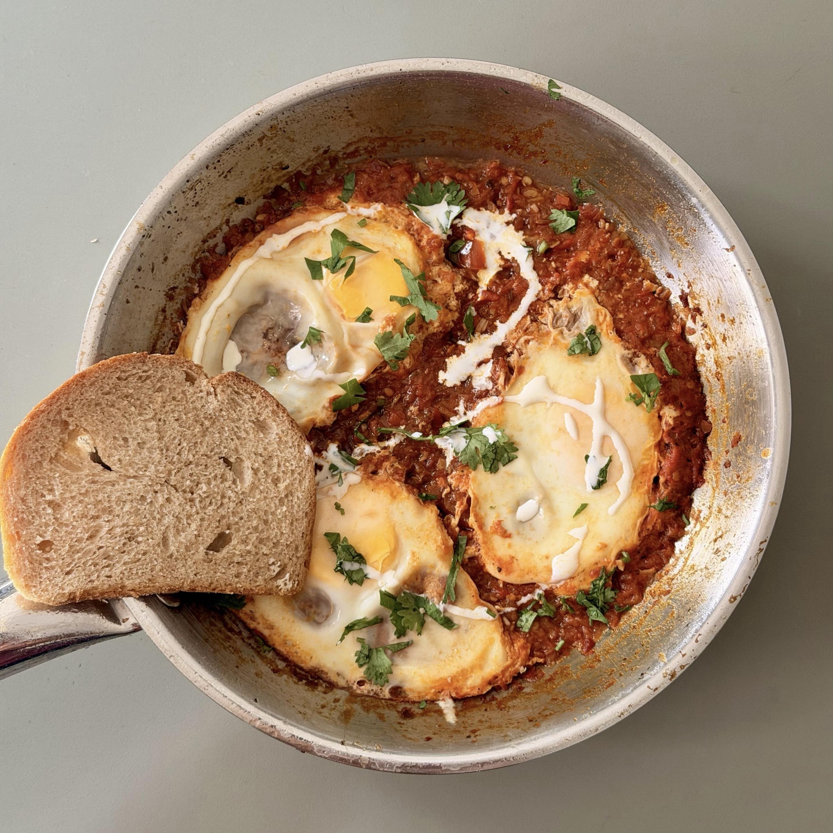 A color, medium-angle photograph in square aspect ratio that captures a homemade skillet of shakshouka resting on a smooth, neutral white countertop. The dish is served directly in a round, well-used stainless steel frying pan, filled with a rich, textured, reddish-brown tomato and pepper base. Nestled into the chunky sauce are three poached eggs with soft whites and visible, intact bright yellow yolks. Drizzled across the top of the dish are swirls of creamy white sauce, garnished with finely chopped cilantro leaves sprinkled throughout. Positioned on the left side, resting partly over the metallic rim of the pan and the sauce, is a single thick slice of crusty whole-grain bread with a visible porous interior texture. The pan's metallic handle extends off to the lower-left edge.