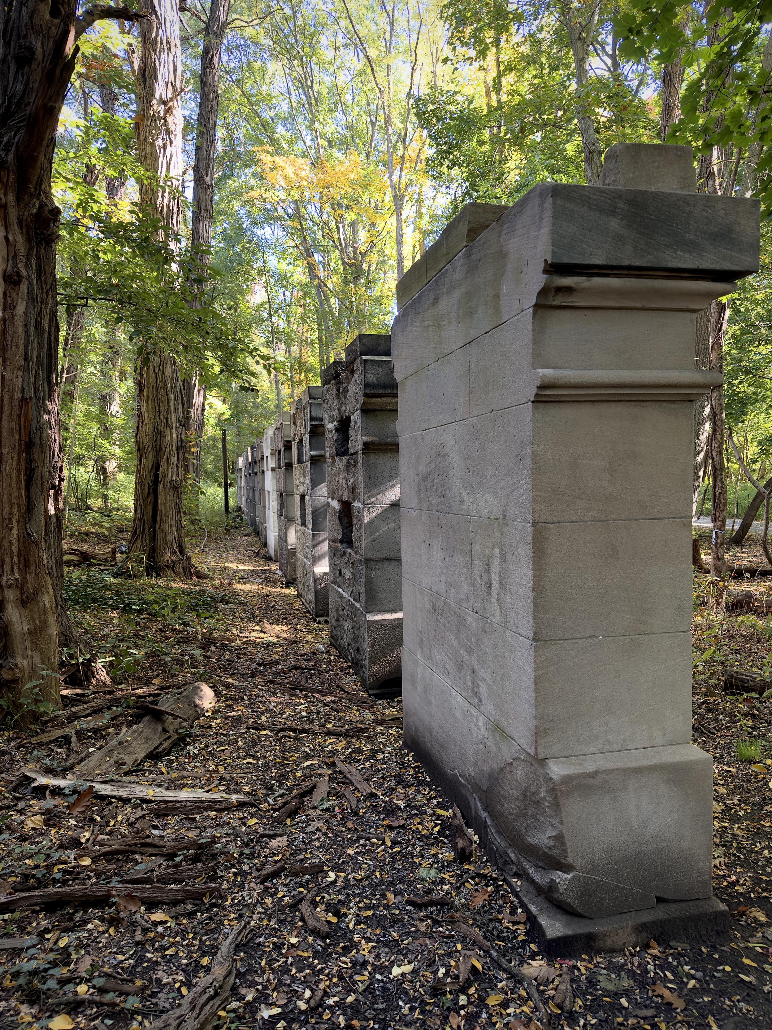 A color, wide-angle photograph in portrait orientation that captures the ‘Thirteen Stone Pillars’ landmark in Van Cortlandt Park, the Bronx, New York during the morning time. The scene shows a line of thick, rectangular stone slabs standing in a forest setting. The stone structures extend in a straight line from the right foreground toward the background, receding into the center-left distance. The prominent pillar in the immediate foreground features smooth, light gray stone blocks stacked vertically, topped with decorative architectural moldings. Further down the row, the subsequent stones vary in color and texture, ranging from darker rough-hewn granite to lighter dressed stone, illustrating different masonry samples. The scene is framed by a dense woodland with tall, slender tree trunks on the left and right, their leafy canopy creating dappled sunlight across the ground. Green foliage dominates the forest background, interspersed with hints of yellow autumn leaves overhead. The ground along the path is covered with fallen brown leaves, small twigs, and dry wood debris, giving the natural environment a quiet, wooded atmosphere.