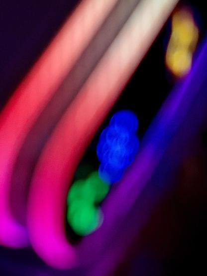A color, macro photograph in portrait orientation that captures vibrant, heavily out-of-focus neon lights set against a dark background. Smooth, sweeping bands of light arch diagonally across the frame, transitioning through glowing shades of magenta, pink, and pale peach. The soft, blurred quality of these neon tubes gives them a smooth, liquid-like appearance, creating a bold sweep of color that dominates the left and upper portions of the image. Nestled within the dark space formed by the curving neon bands are overlapping, circular bokeh orb patterns in vivid hues. Small clusters of bright blue and vibrant green light orbs sit near the center, while a warm golden-yellow bokeh glow shines softly in the upper right background. Deep purple light sweeps along the lower-right edge, framing the dark background and accentuating the luminous, dreamlike atmosphere of the nighttime street scene.