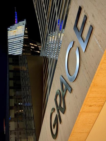 A color, wide-angle photograph in portrait orientation that captures the striking modern architecture of skyscrapers in Midtown Manhattan. Occupying the right half of the frame in strong perspective is the Grace Building, featuring a beige, textured stone facade angled dramatically upward into the clear black night sky. Dimensional, dark metallic lettering spelling out "GRACE" is mounted diagonally along the beige surface. Next to it, the building's sweeping glass curtain wall curves concave-upward, reflecting illuminated window bands and vertical architectural lines from the surrounding city lights. In the background to the left, Bank of America Tower towers over the lower glass structure beside the Grace Building. Its prominent sloped, angular glass crown and spire are brightly illuminated against the dark sky, casting a radiant white and blue glow. The surrounding dark skyscraper facades reflect geometric grid patterns of glowing office windows, creating a sleek, high-contrast urban composition of metal, stone, glass, and light.