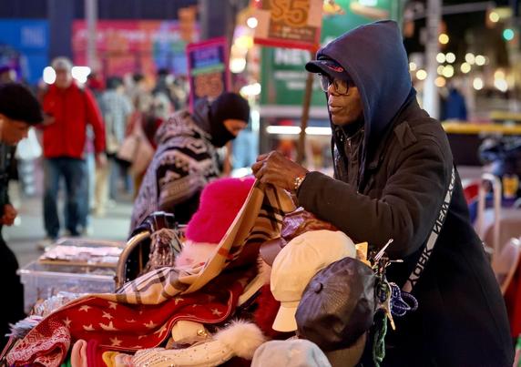A color, wide-angle photograph that captures a close-up, night scene of a street vendor managing his display table along 42nd Street in the Times Square neighborhood of Manhattan. The primary subject is a dark-skinned man wearing a blue-hooded jacket over a dark coat, a baseball cap, and glasses, leaning forward over a dense pile of merchandise. His hands are displaying items to a customer over a table packed with various winter accessories and goods, including plaid scarves, red patterned winter wraps, fuzzy beanies, white and brown caps, and small souvenirs in plastic storage bins nearby. He carries a black shoulder strap featuring white block lettering that reads "NEW YORK". In the immediate background, another individual dressed in a patterned sweater and black face covering is standing behind the display, assisting with or looking through the merchandise. Beyond the vendor table, the bustling urban environment of Times Square is softly blurred, showing pedestrians walking by in warm jackets, bright city signage displaying price tags like "$5," and glowing yellow street and traffic lights reflecting off the night pavement. The warm lighting highlights the textured fabrics of the winter clothing against the dark night atmosphere of the city.