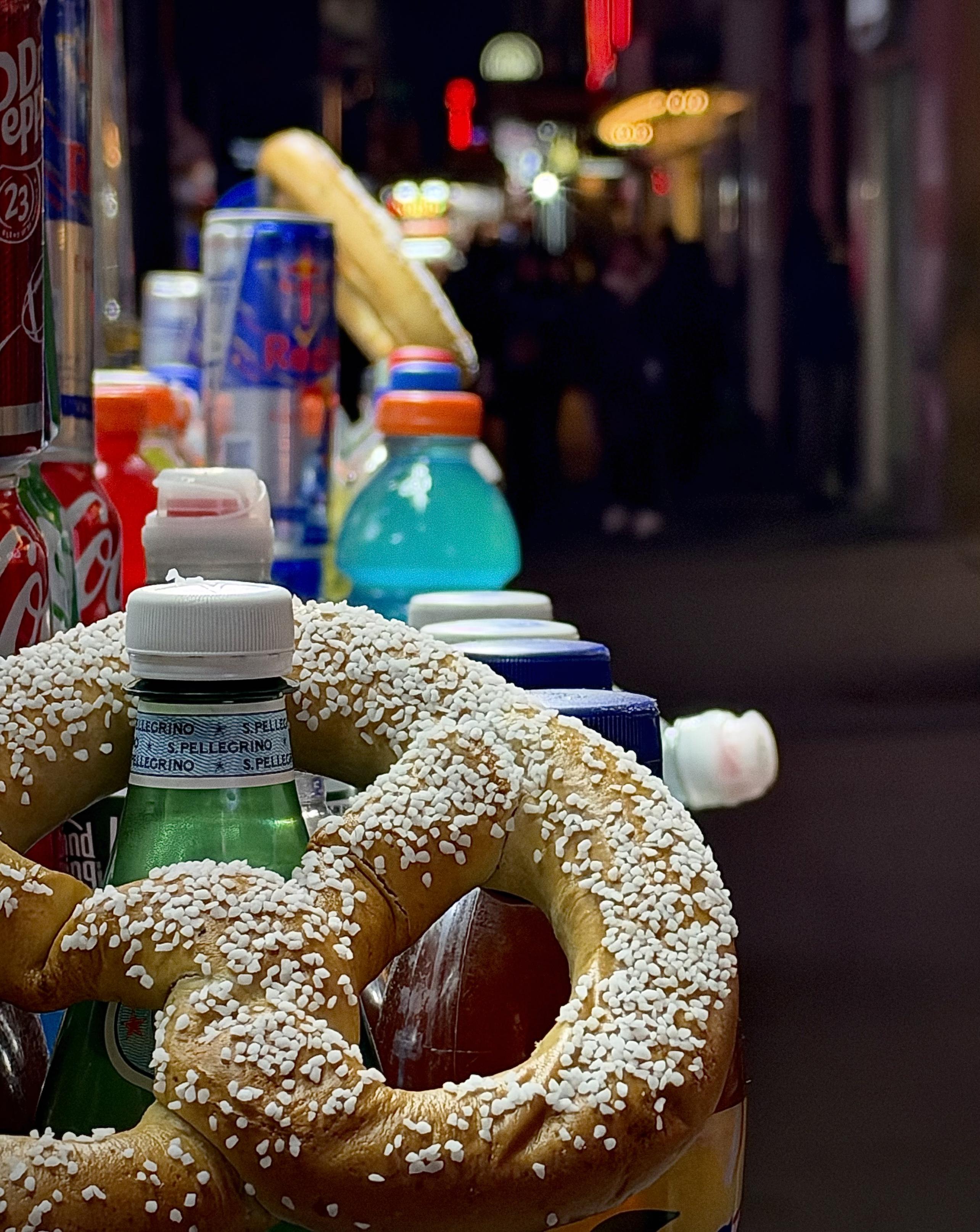 A color, wide-angle photograph in portrait orientation that captures the vibrant display of a street vendor cart at night in New York City's Times Square. In the foreground, a large, golden-brown pretzel generously coated in coarse white salt loops around green bottles of S.Pellegrino sparkling water. Nestled alongside and behind the pretzel are various canned and bottled beverages, including Dr Pepper, Coca-Cola, Red Bull, and bright blue Gatorade. Higher up on the display stand, a freshly baked baguette rests diagonally above the colorful array of drinks.  The background features a shallow depth of field, with shadowy figures of pedestrians walking along the sidewalk. Glowing yellow streetlights, neon signs, and distant city lights create a warm, blurred bokeh effect against the dark urban atmosphere, emphasizing the vivid textures and bright colors of the street food display in the foreground.